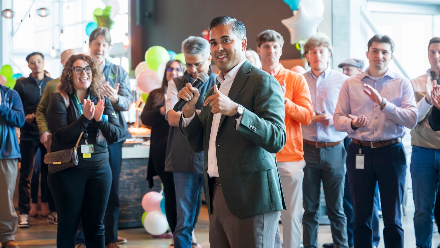 Man in teal blazer gives thumbs up while colleagues applaud at a festive corporate celebration event.