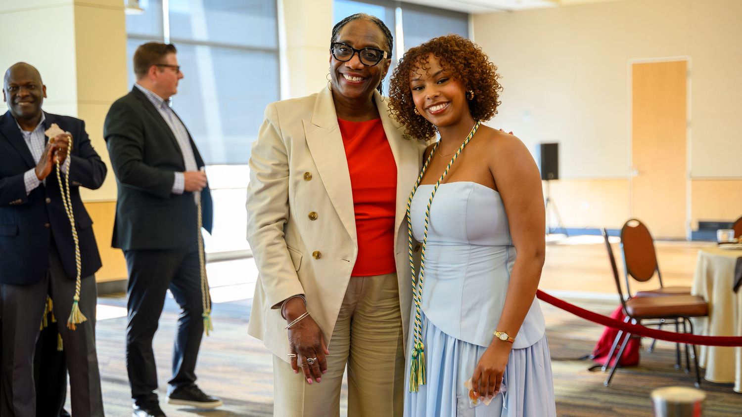 Two elegantly dressed individuals pose together smiling at a formal event, with guests socializing in the background.