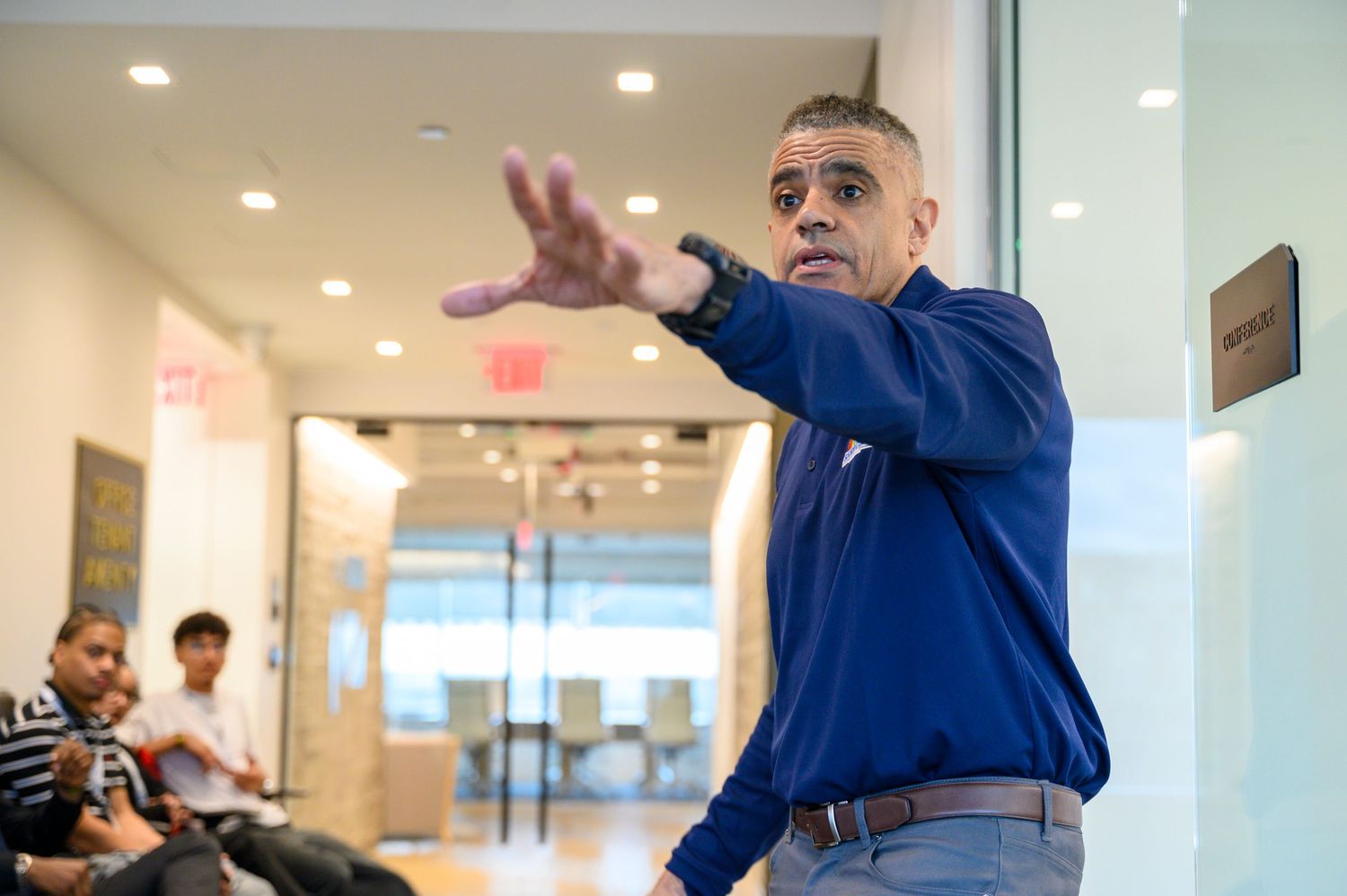 Man in blue shirt gesturing while speaking to a group in a modern office hallway setting.