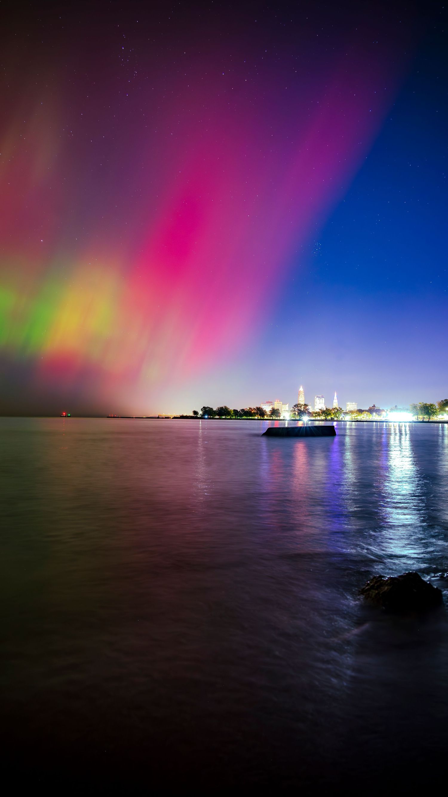 Vibrant aurora borealis with pink and green hues reflecting over calm water near a city skyline at night.