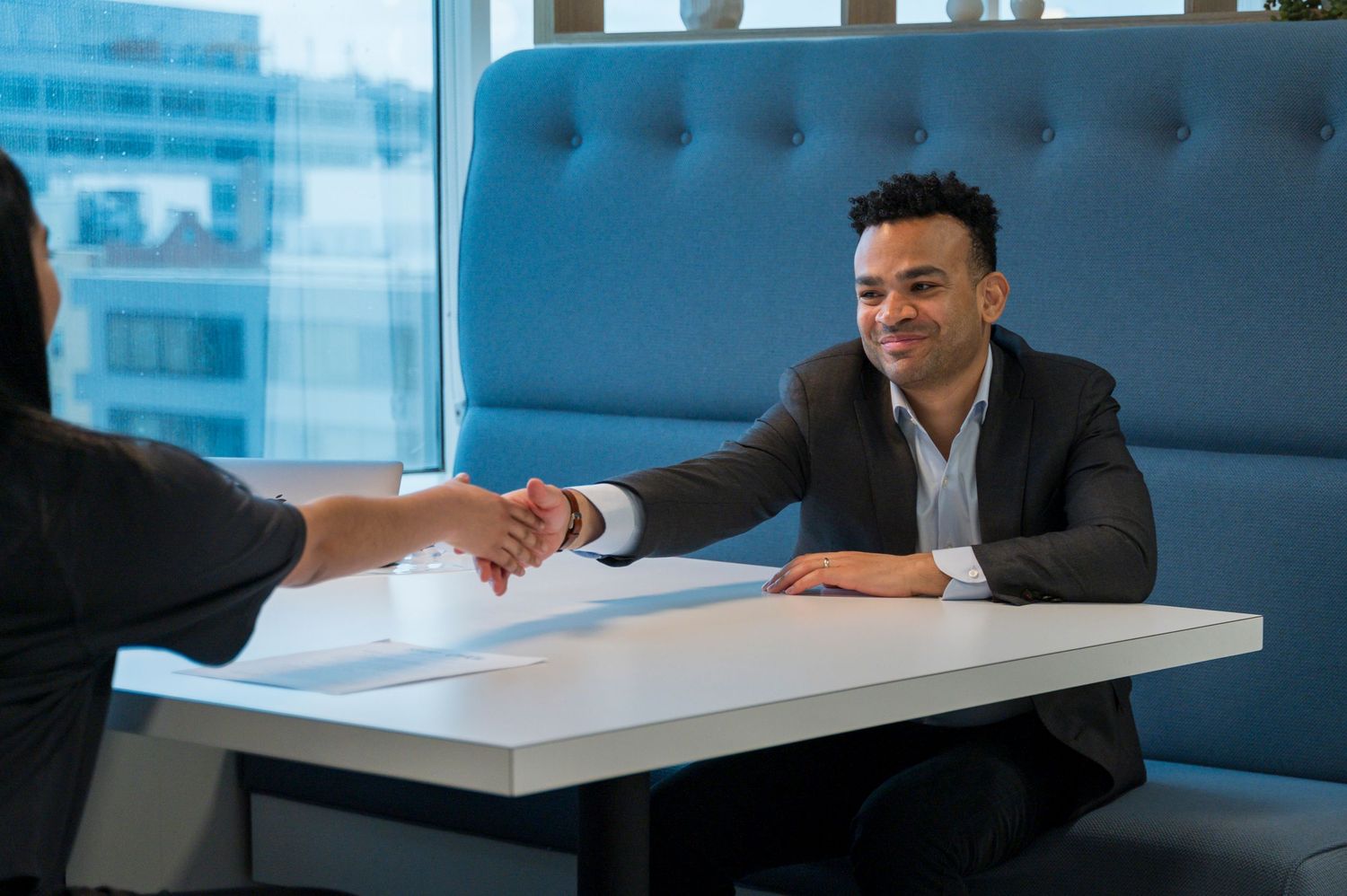 Professional man in suit smiling and shaking hands across a white table in a modern office setting.