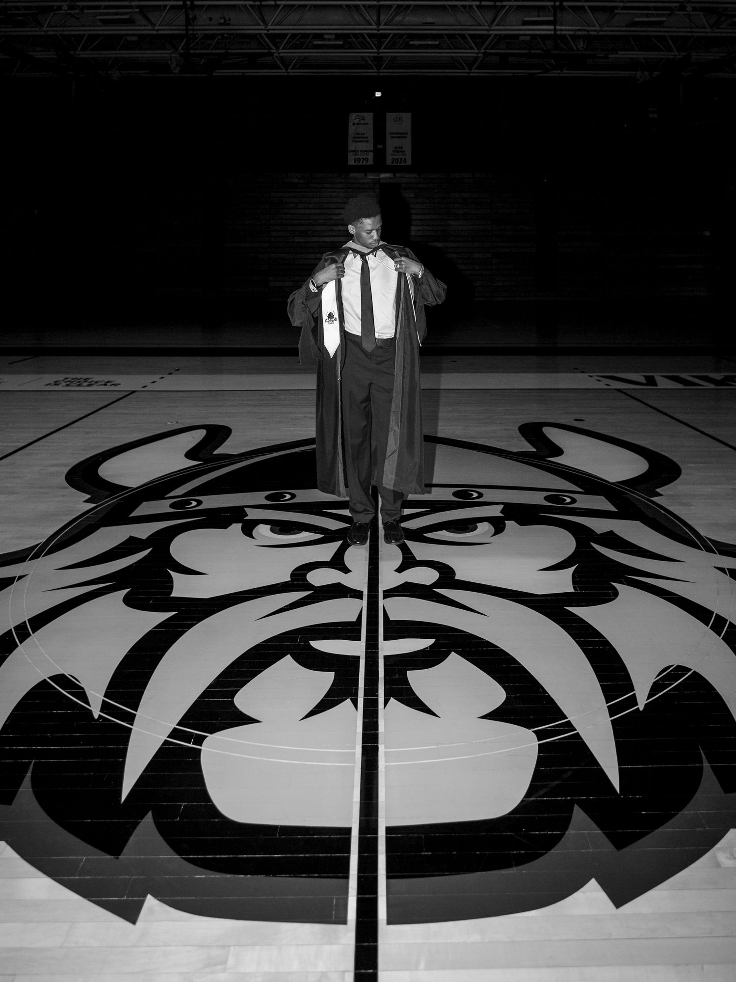 Black and white photo of a graduate in cap and gown standing on a basketball court center logo.