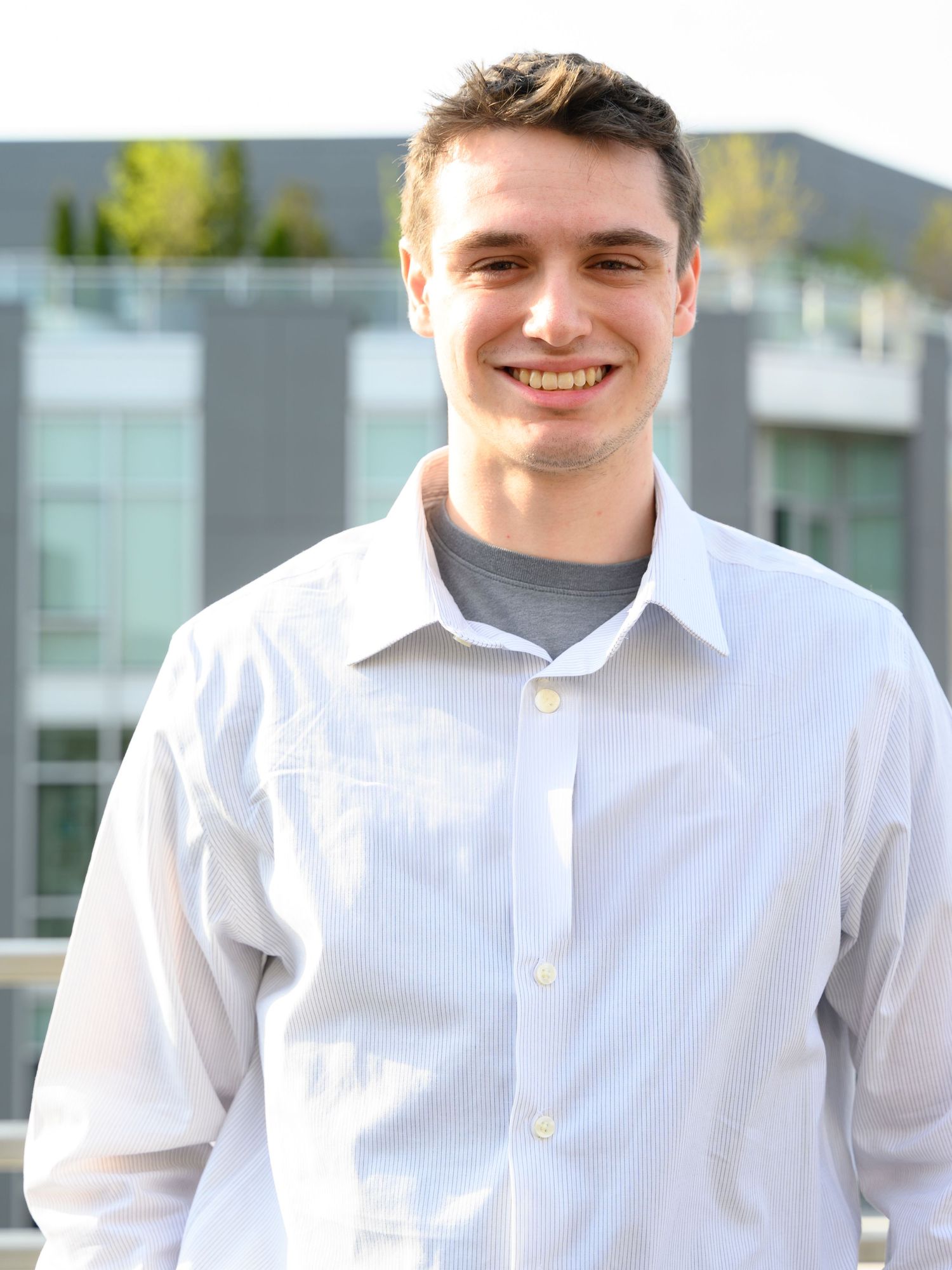 Smiling young man in white button-down shirt standing outdoors near a modern building.