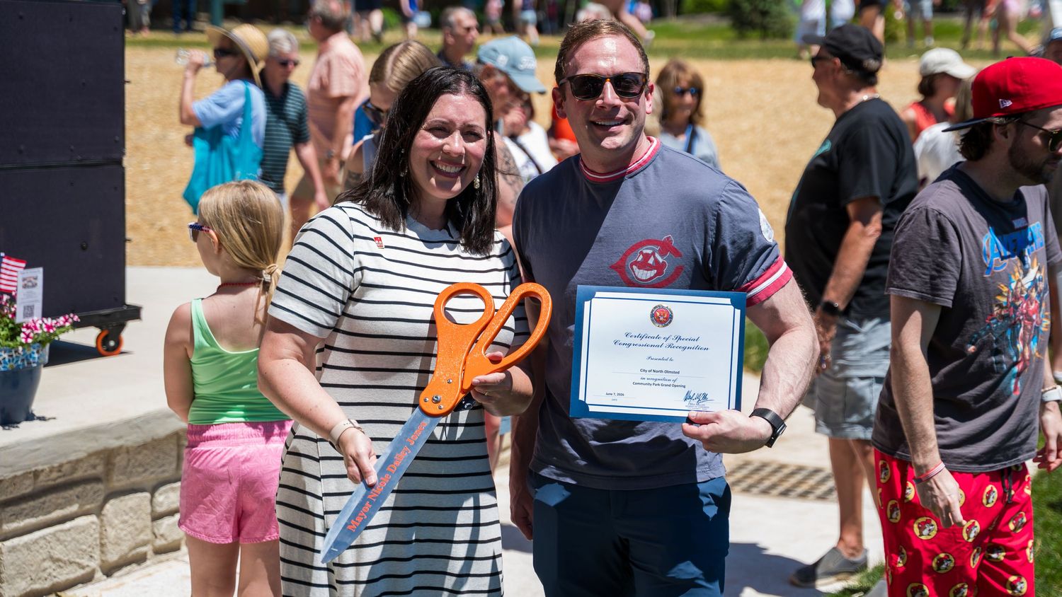 Two people smile holding oversized scissors and a certificate at an outdoor ribbon-cutting ceremony on a sunny day.