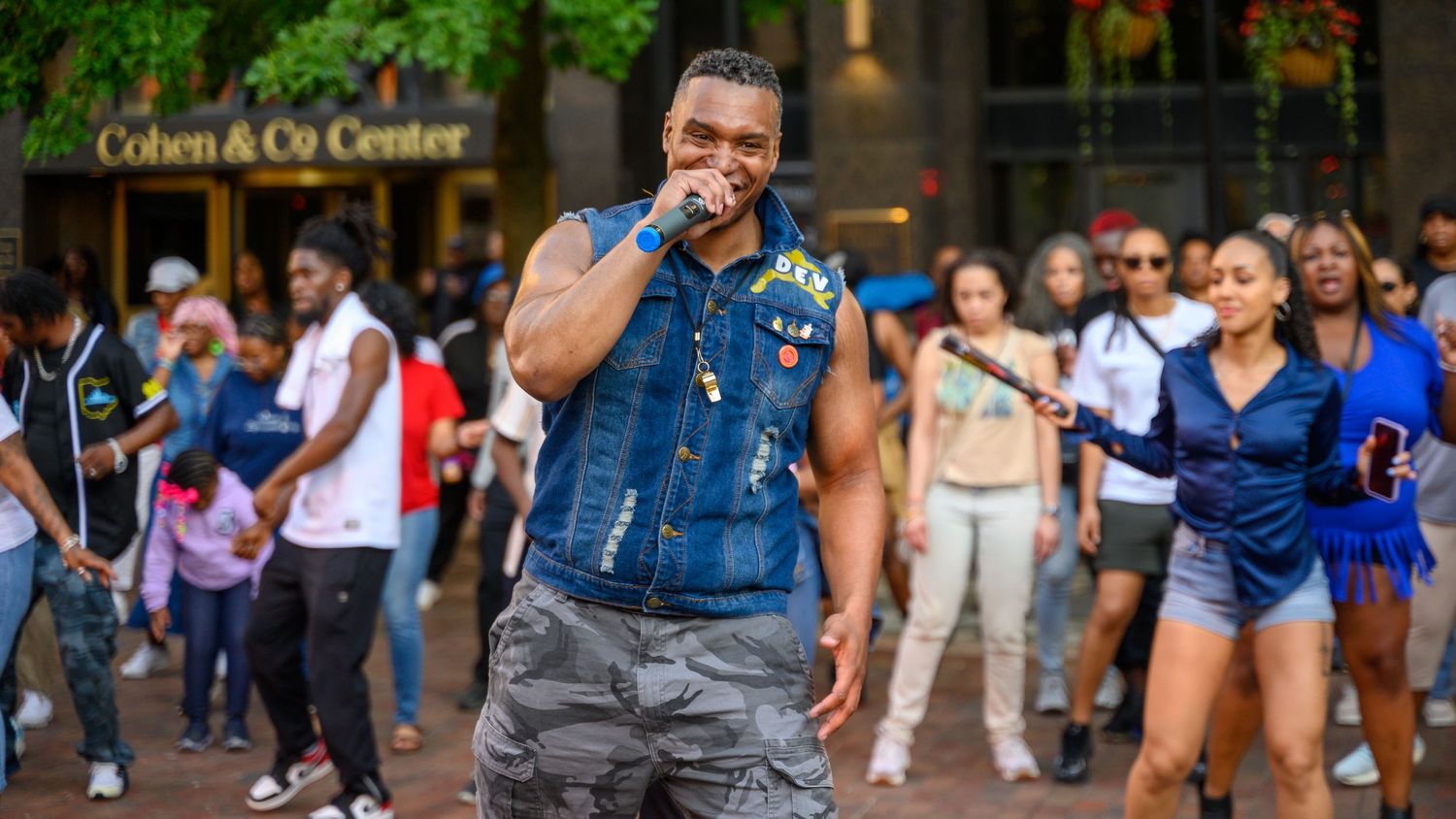 A man performs outdoors at Cohen & Co Center, singing into a mic wearing a denim vest, with a crowd behind him.