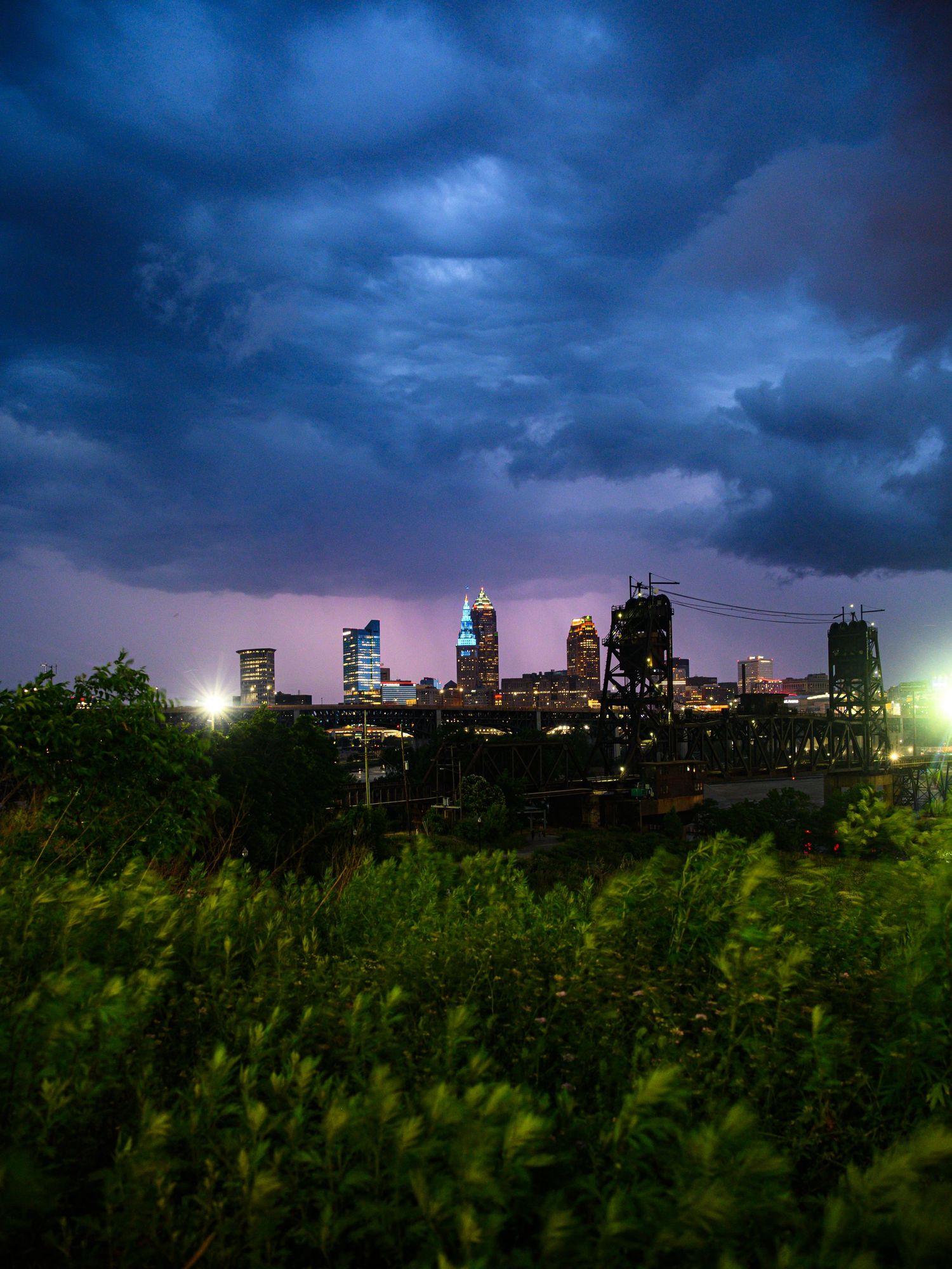 Dramatic stormy night skyline with lightning illuminating dark blue clouds over a city with lush green foreground vegetation.