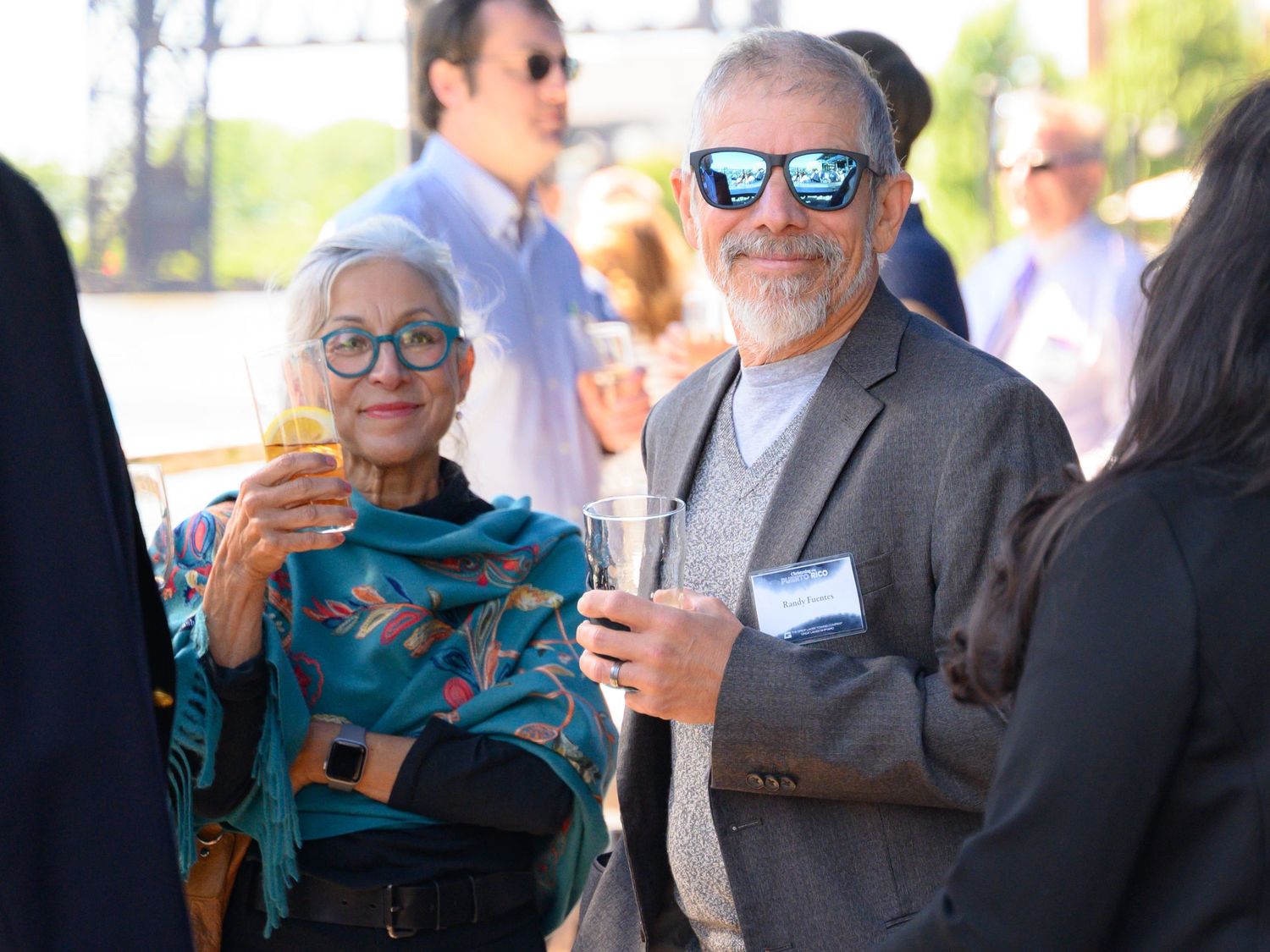Two guests enjoying drinks at an outdoor social event, woman in teal scarf and man in sunglasses holding champagne.