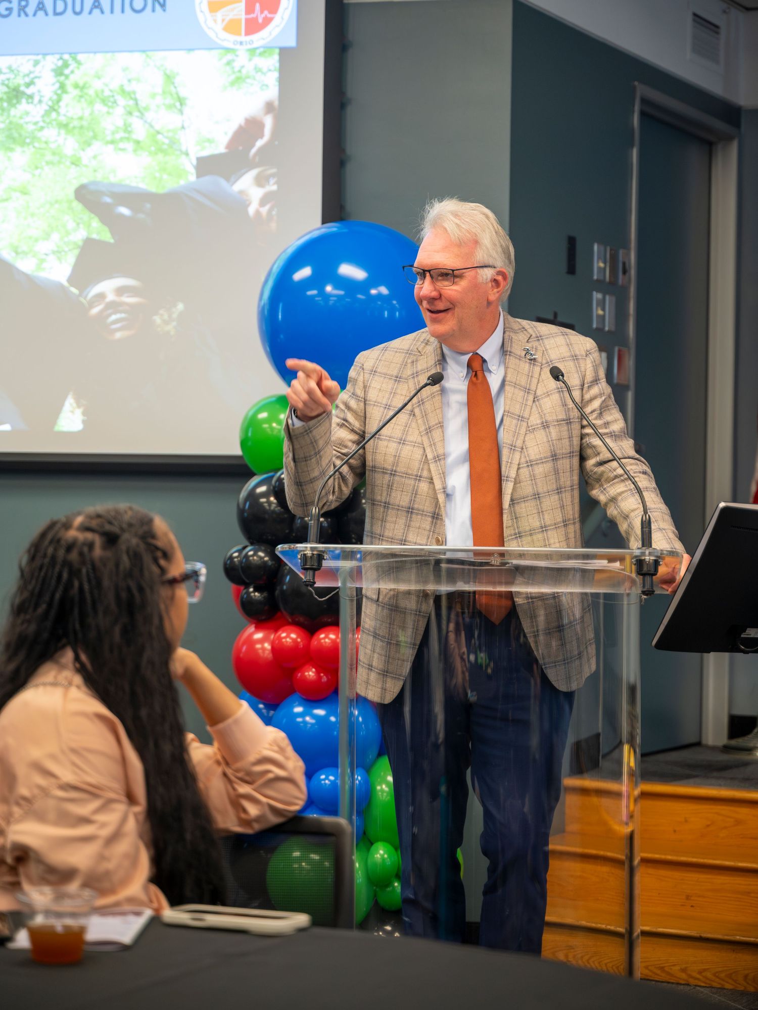 Speaker in plaid blazer addresses audience at podium during graduation event with colorful balloons.