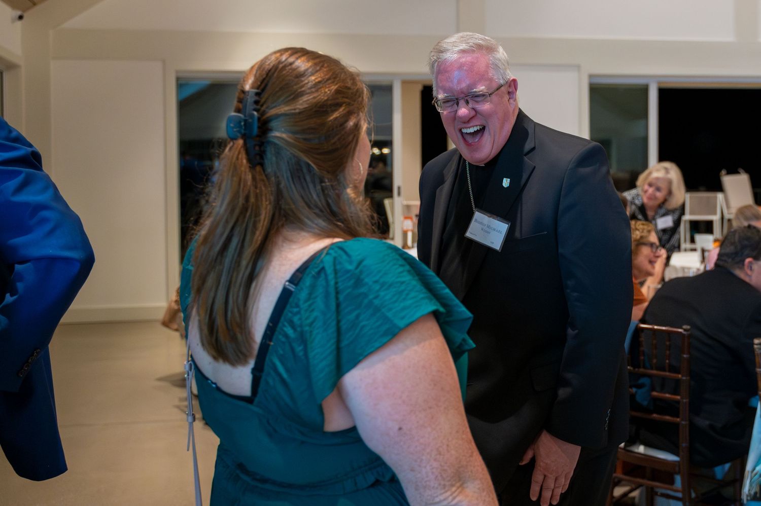 Man with name badge laughing warmly with a woman in teal dress at indoor gala event.