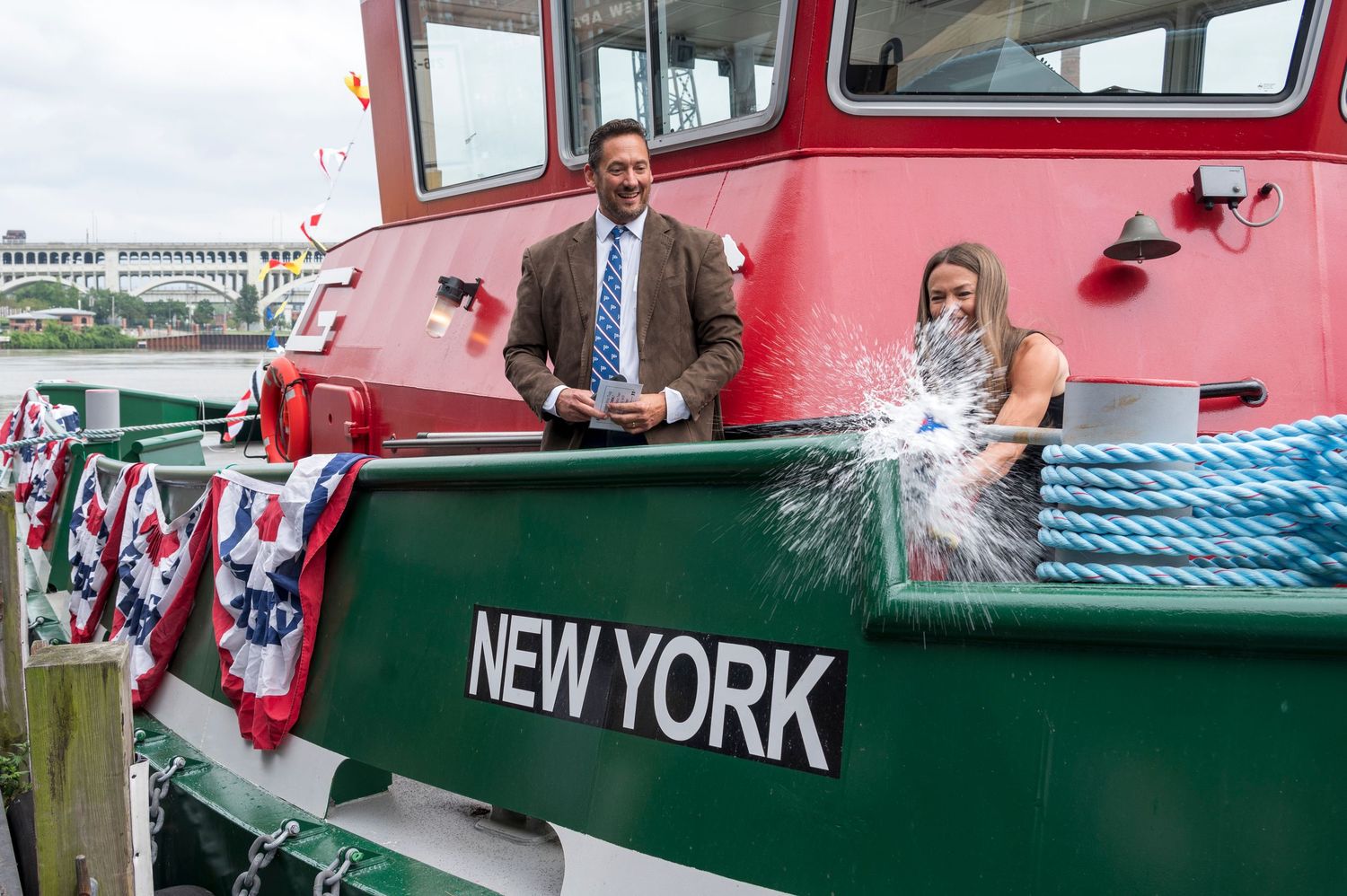 Two people christen a red and green tugboat named New York with champagne at a waterfront ceremony.