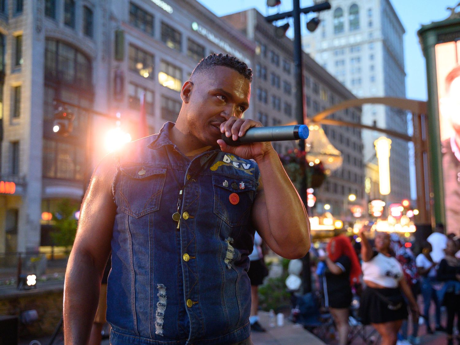 A male rapper performs outdoors at a city street event, singing into a microphone wearing a denim vest at dusk.
