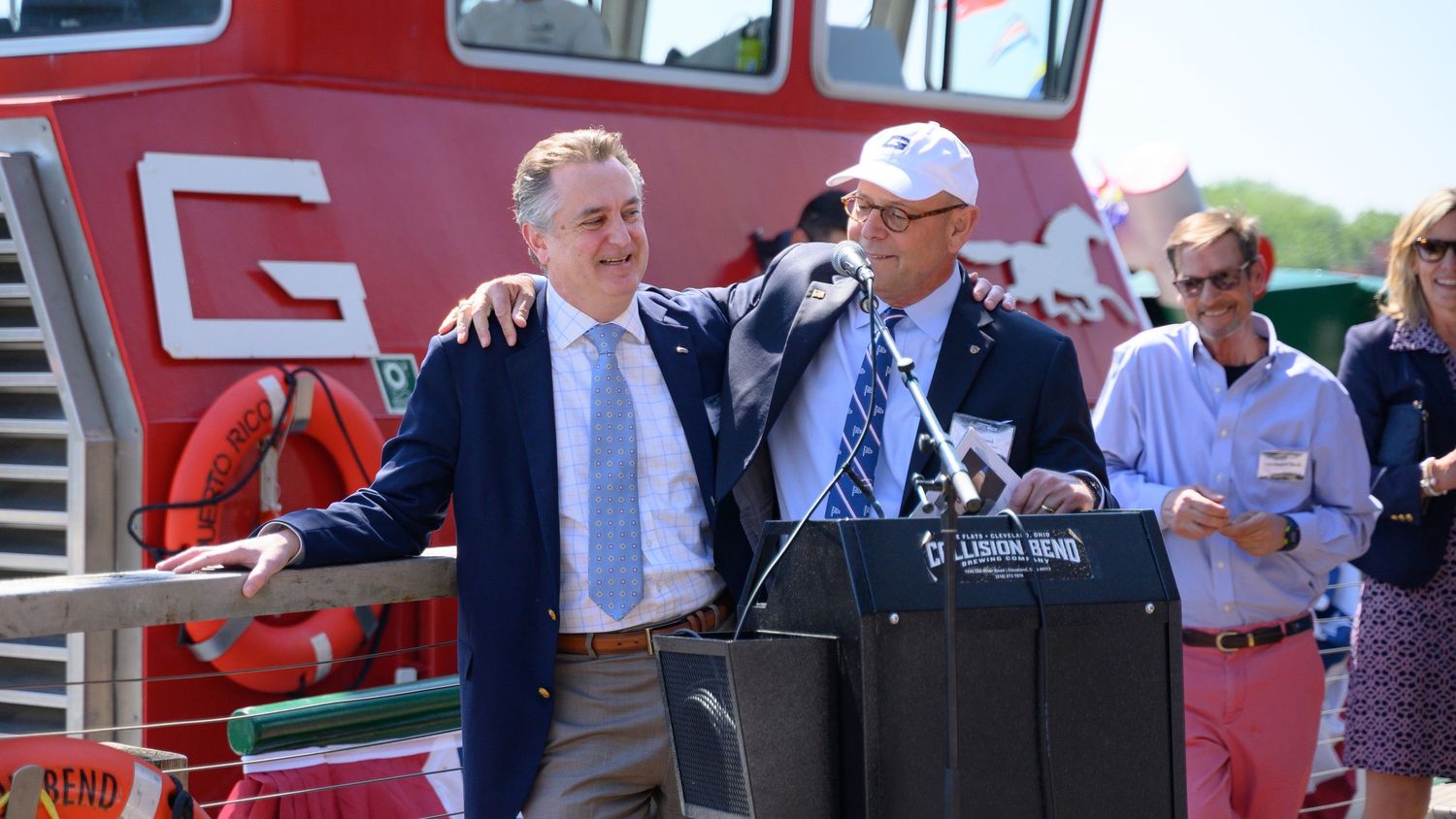 Two men stand at a podium on a red boat during an outdoor event, with guests visible in the background.