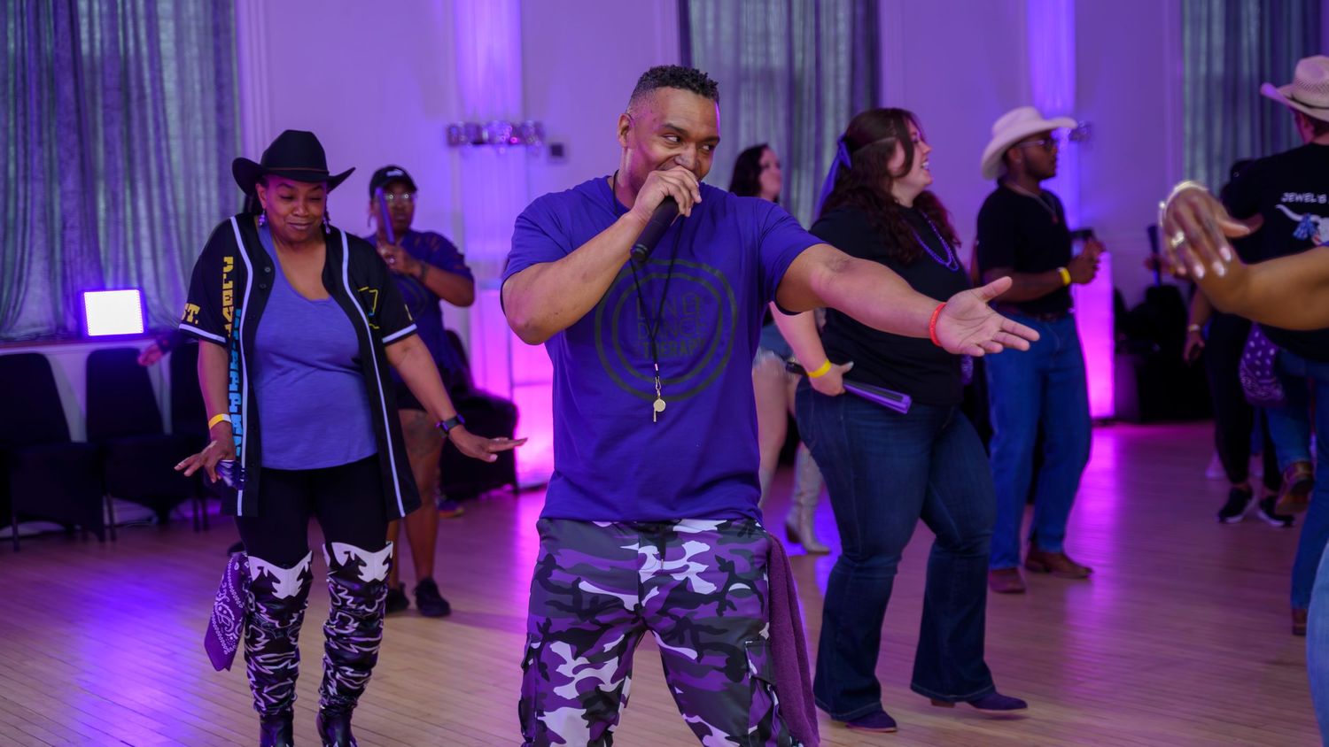 Man in purple shirt leads a line dance at an indoor event with purple lighting.