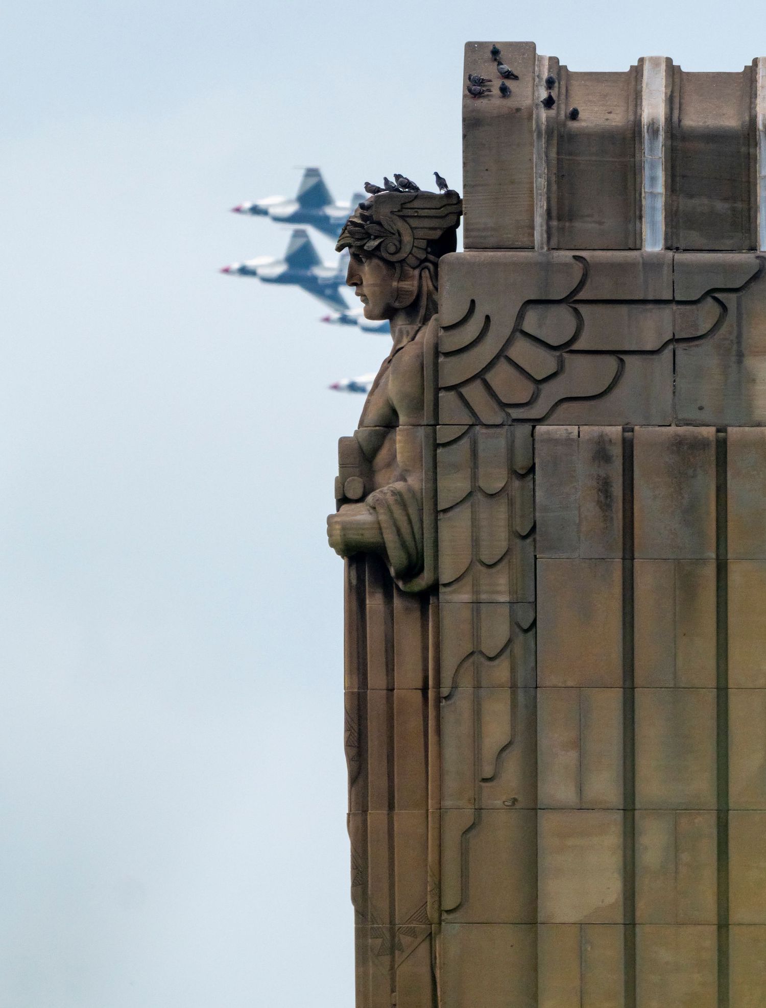 Military jets fly in formation past an Art Deco winged guardian statue on a stone building facade.