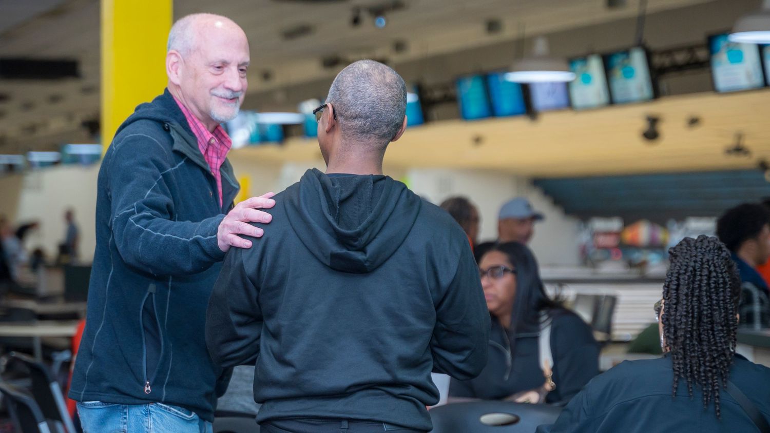Two men engage in friendly conversation at a busy bowling alley.