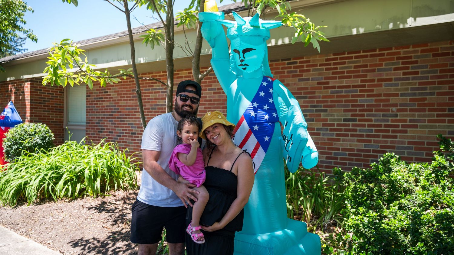 Family poses with Statue of Liberty sculpture outside brick building on sunny day.