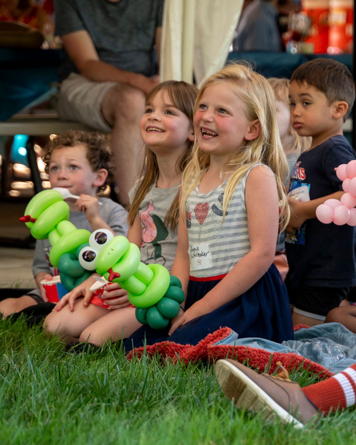 Happy children sitting on grass holding colorful balloon animals at outdoor party event.