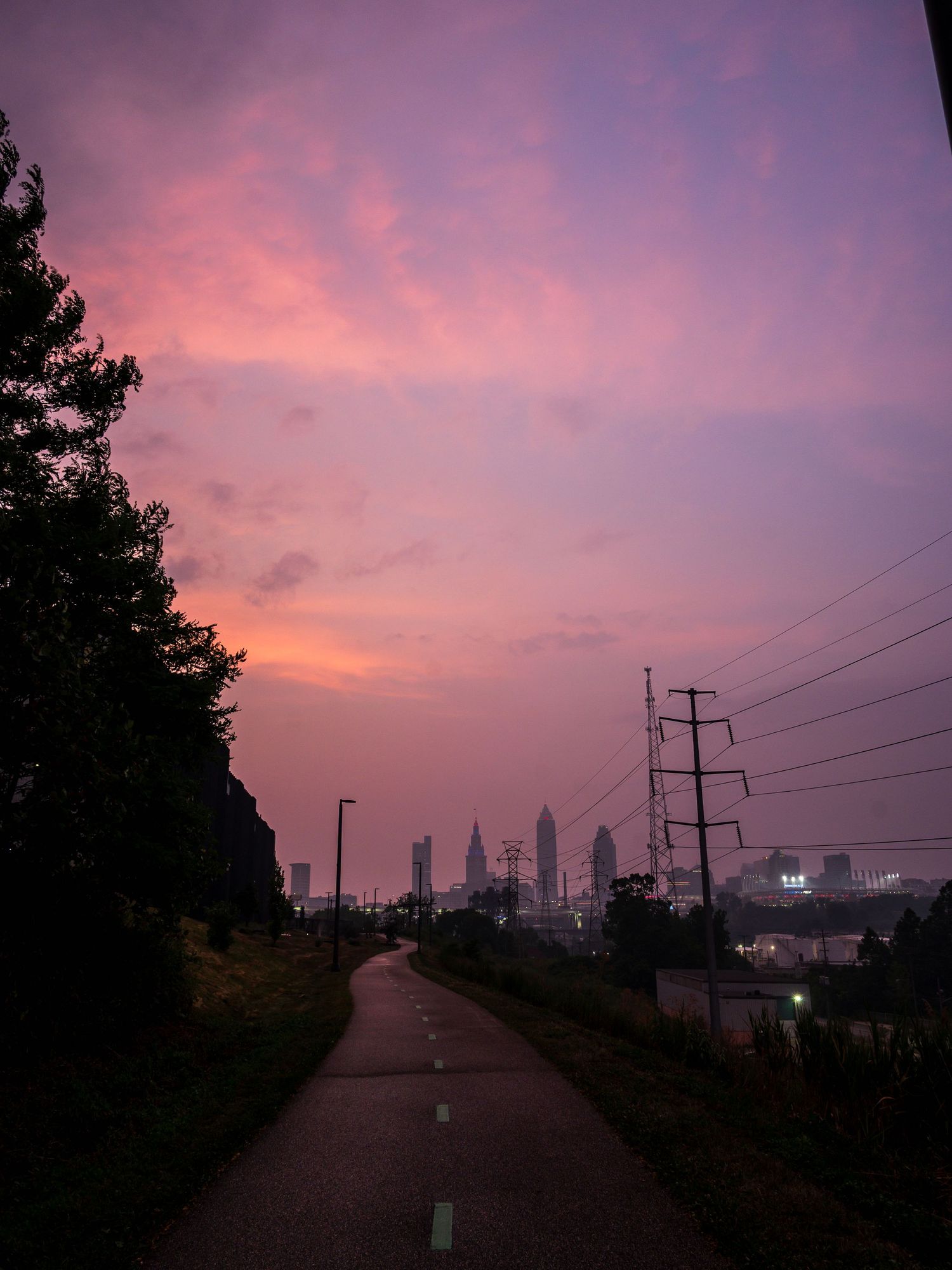 A stunning pink and purple sunset sky over a city skyline viewed from a quiet road flanked by trees and power lines.