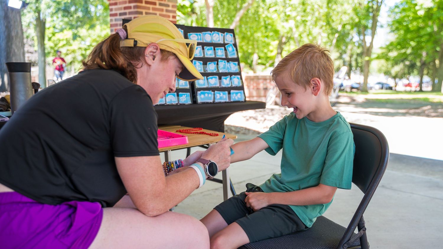 A woman in a yellow cap checks a young boy's arm at an outdoor table during a community health event.