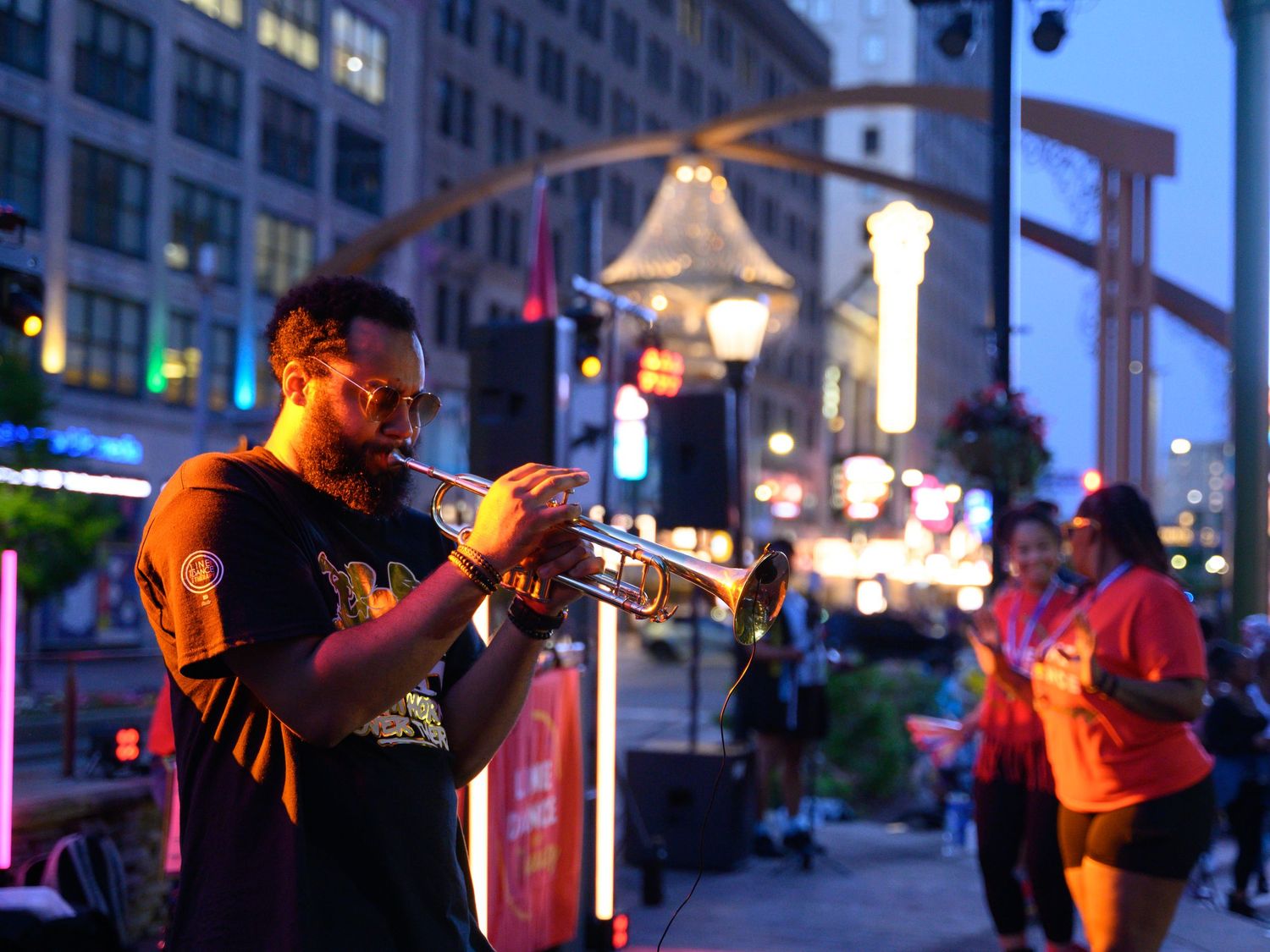 A musician plays trumpet at an outdoor urban street festival at dusk with colorful city lights glowing in the background.