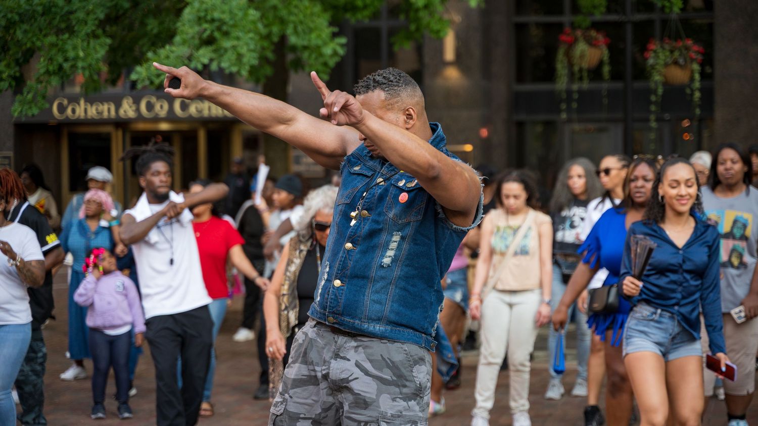 Man in denim vest dabbing while leading a crowd dance in an outdoor urban plaza near Cohen & Co.