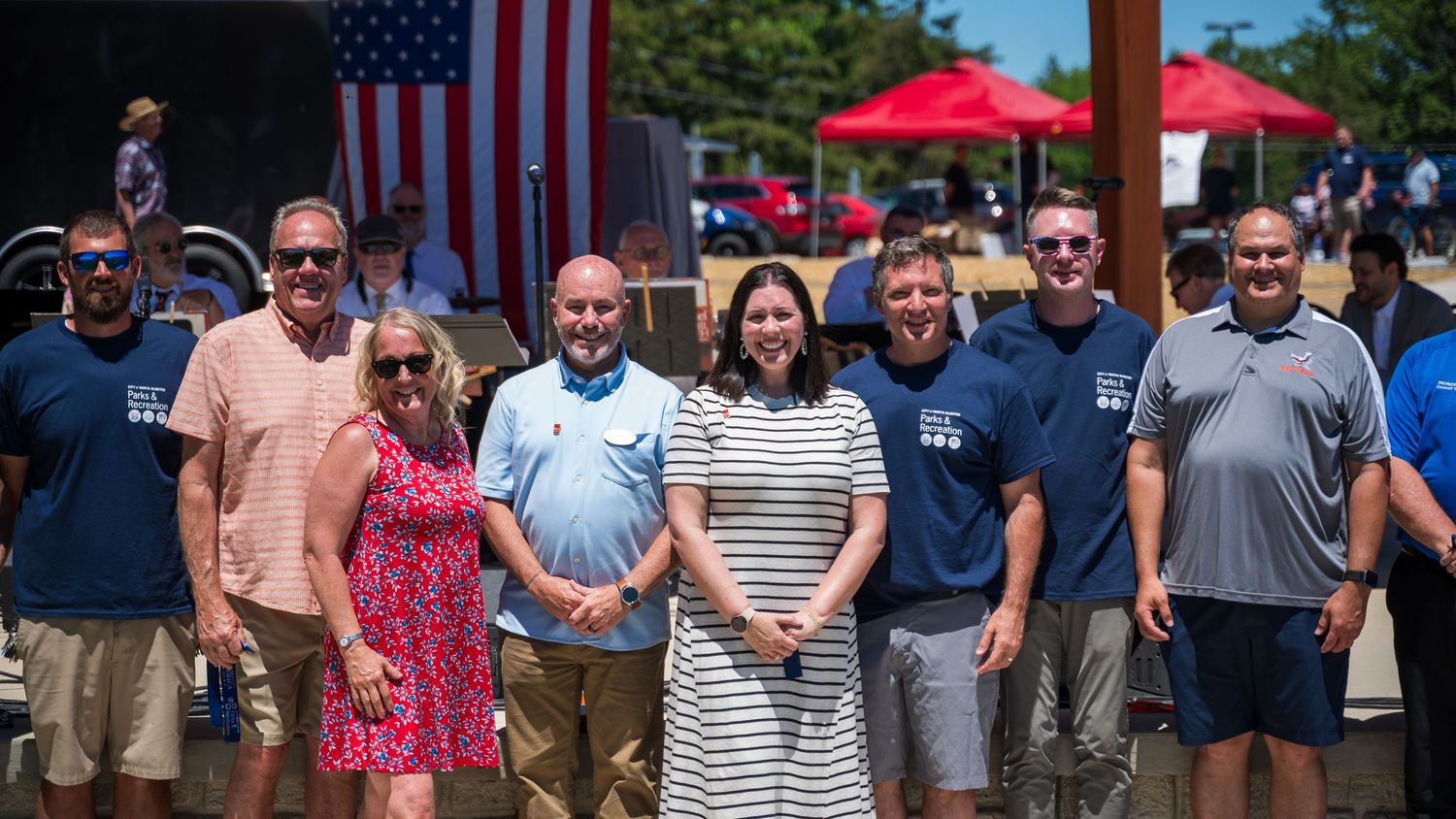 Group of eight people posing outdoors at a patriotic event with American flag and red tent in background.
