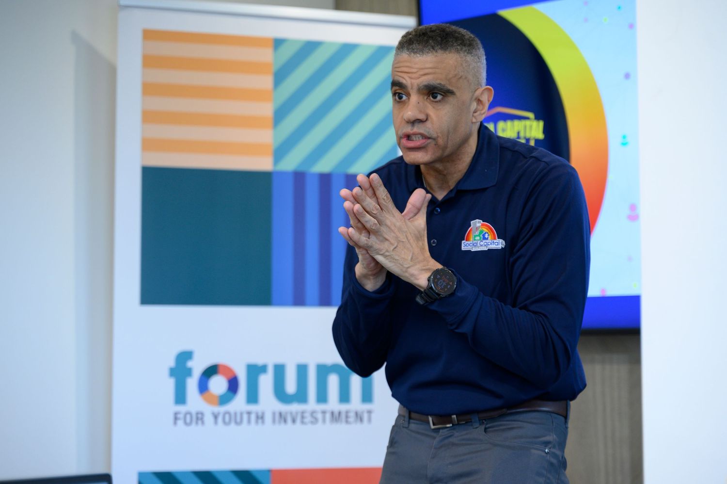 Man speaking at Forum for Youth Investment event, gesturing with hands clasped in front of colorful banner.