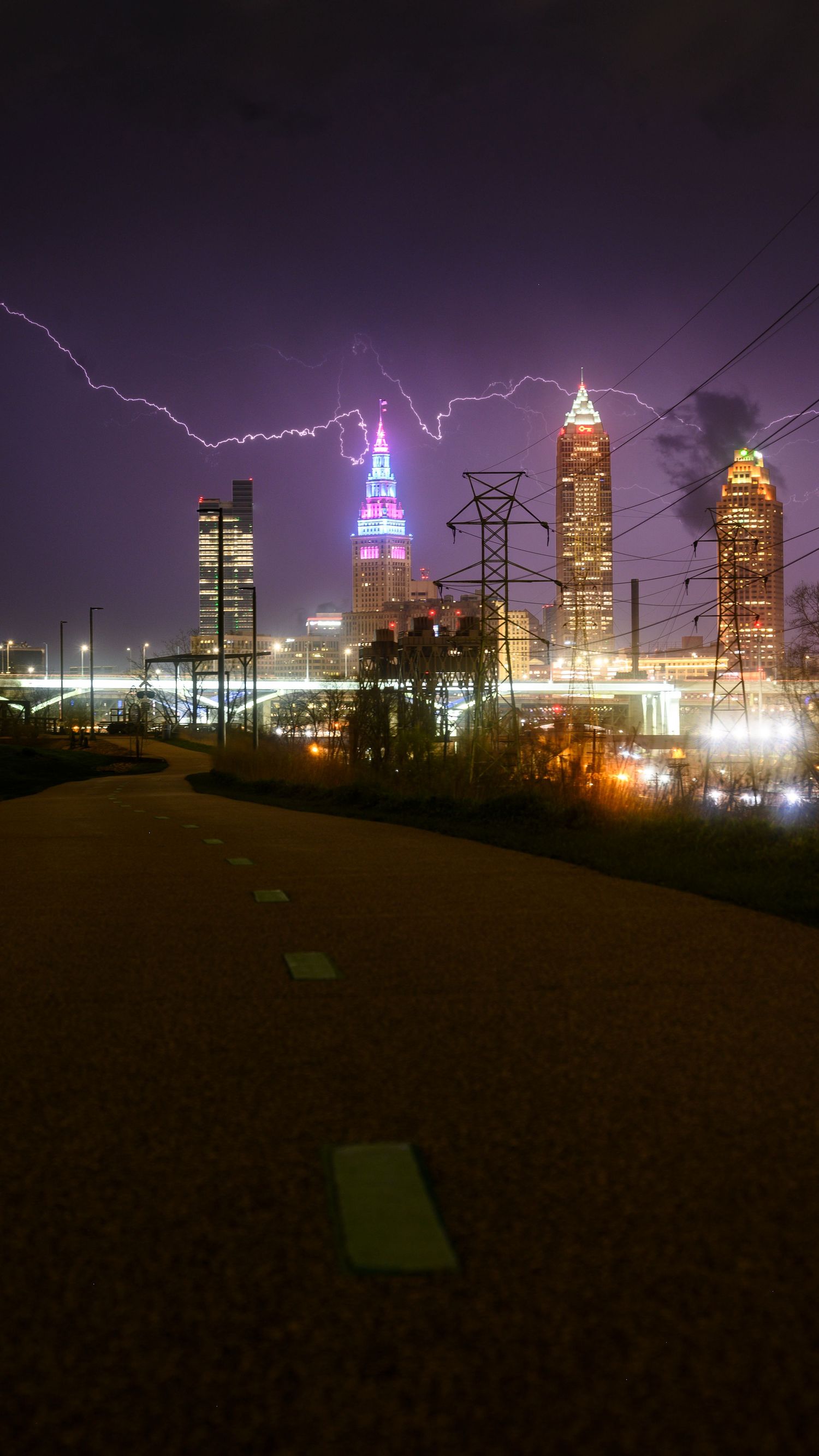 Lightning strikes over illuminated Atlanta skyline at night with colorful skyscrapers and power lines in foreground.