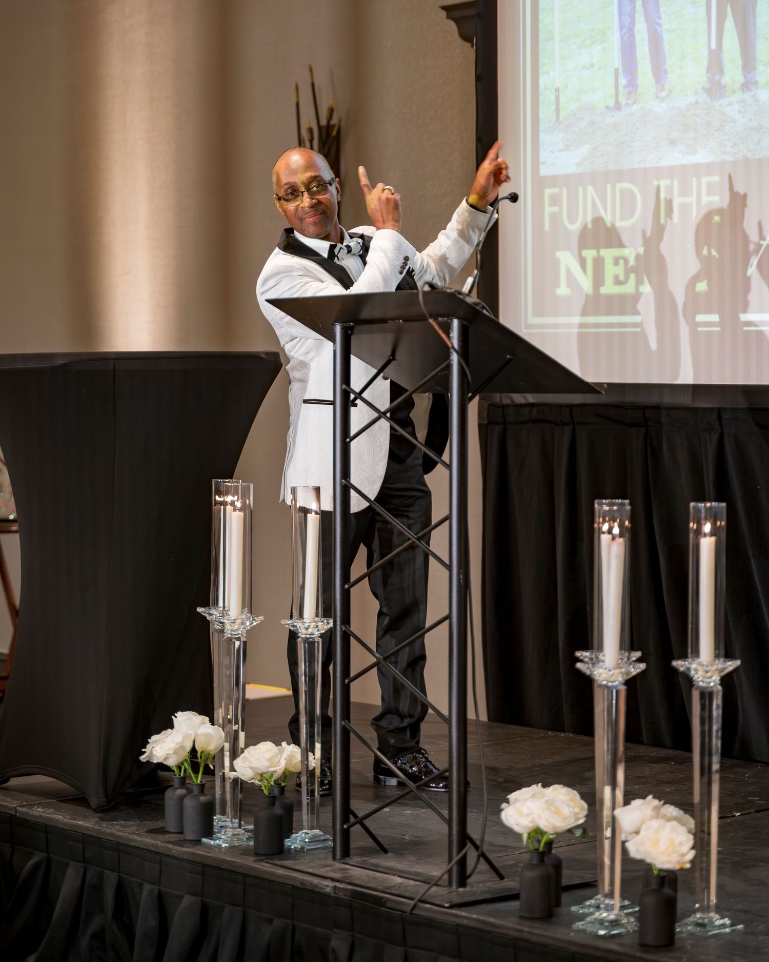 Speaker in white tuxedo gestures at podium during Fund the Next formal fundraising gala event.