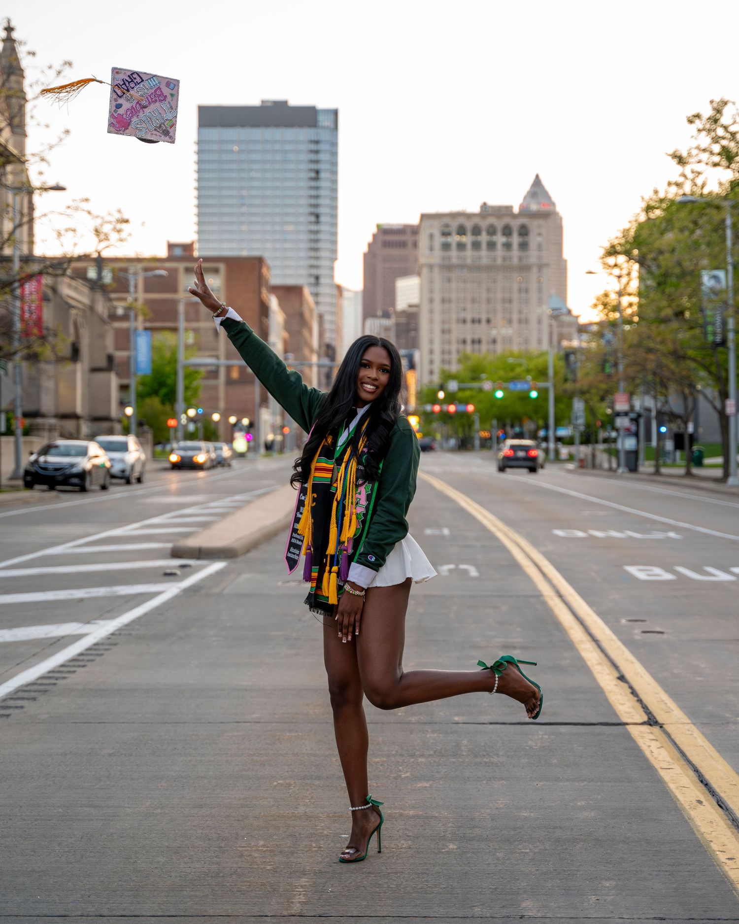 A graduating woman in a green jacket tosses her cap on a city street at golden hour.