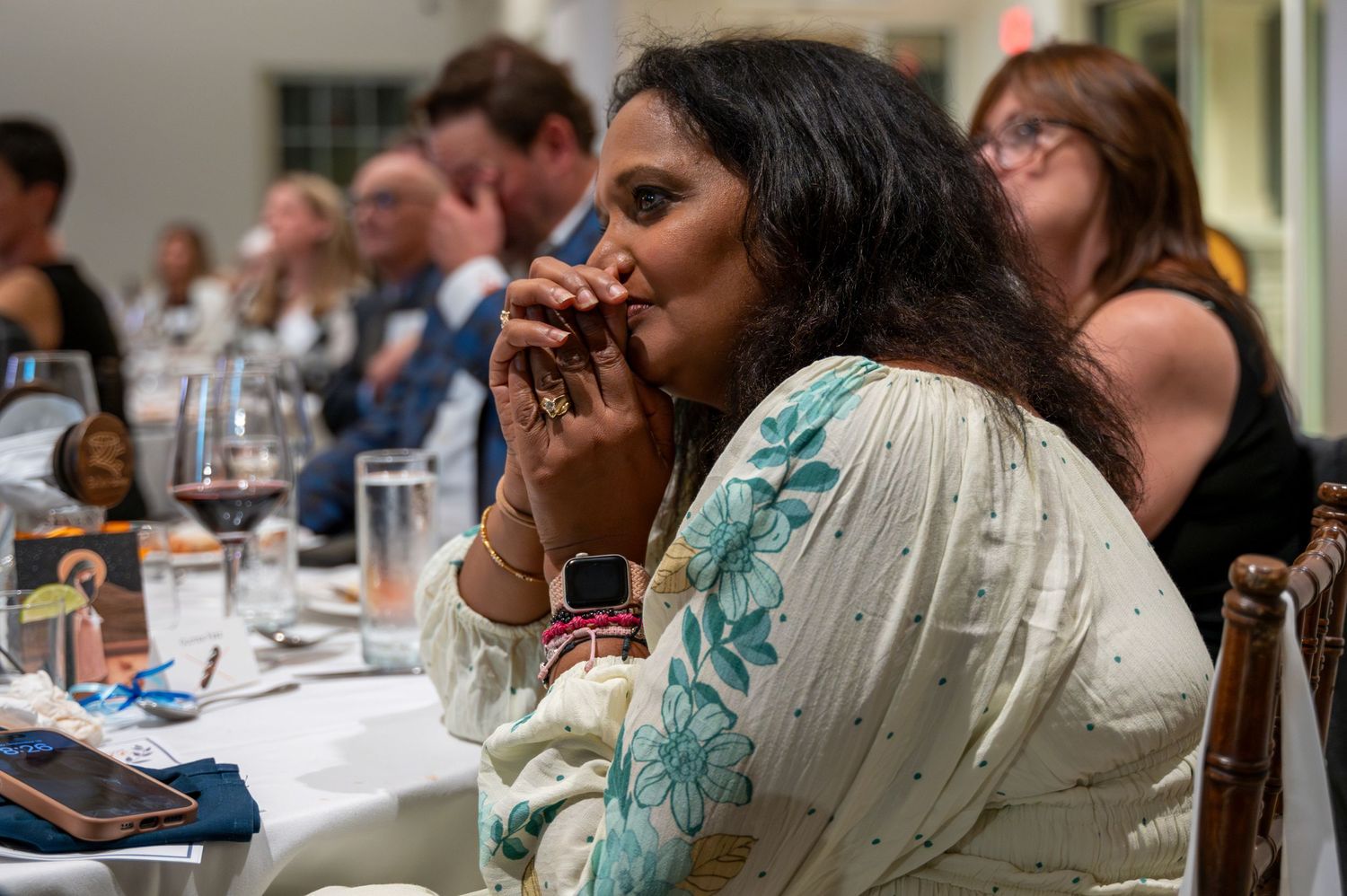 Woman in floral sari listens attentively at an elegant indoor dinner event with wine glasses.