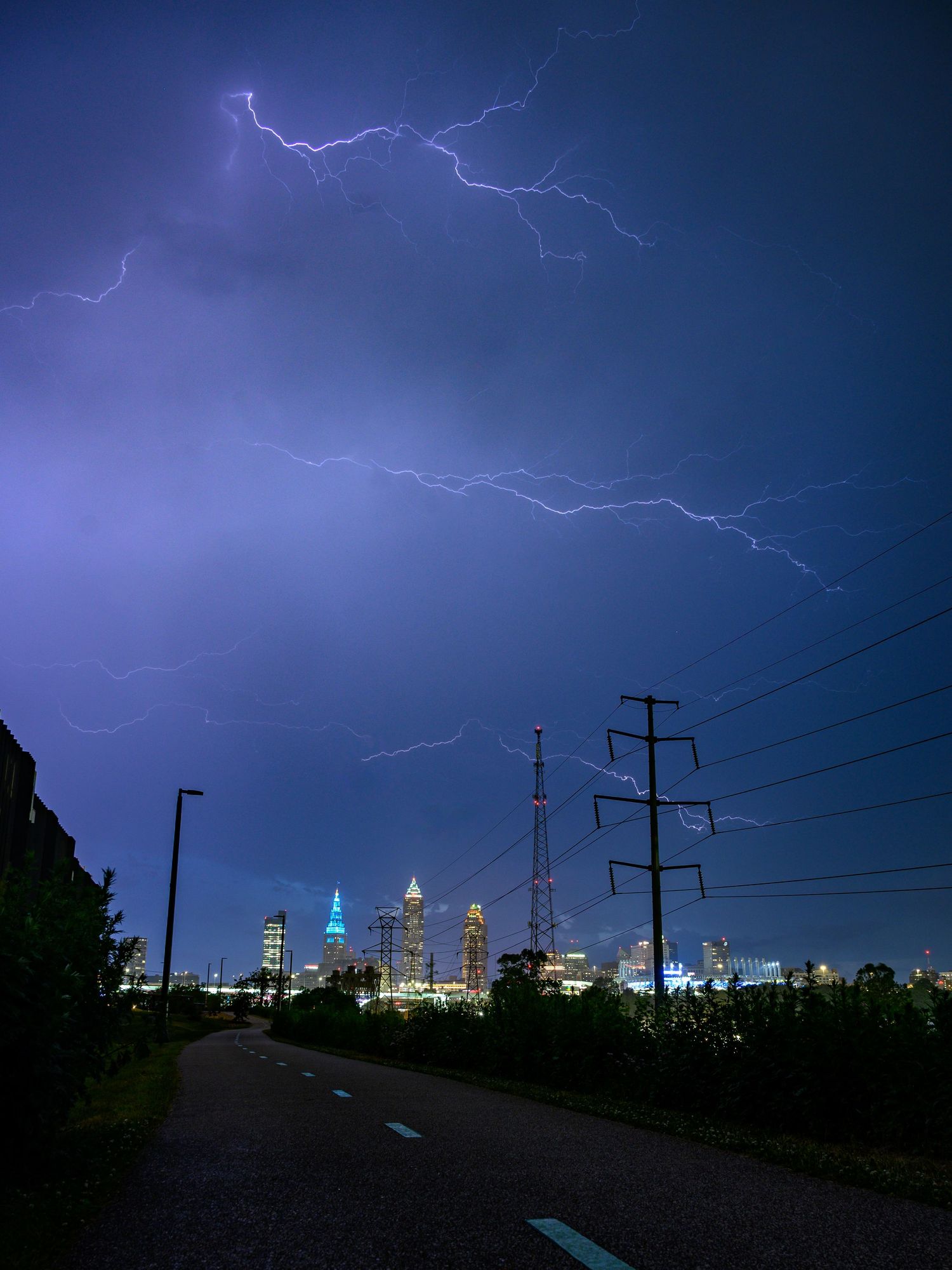 Lightning storm illuminates city skyline at night with power lines silhouetted against purple sky.