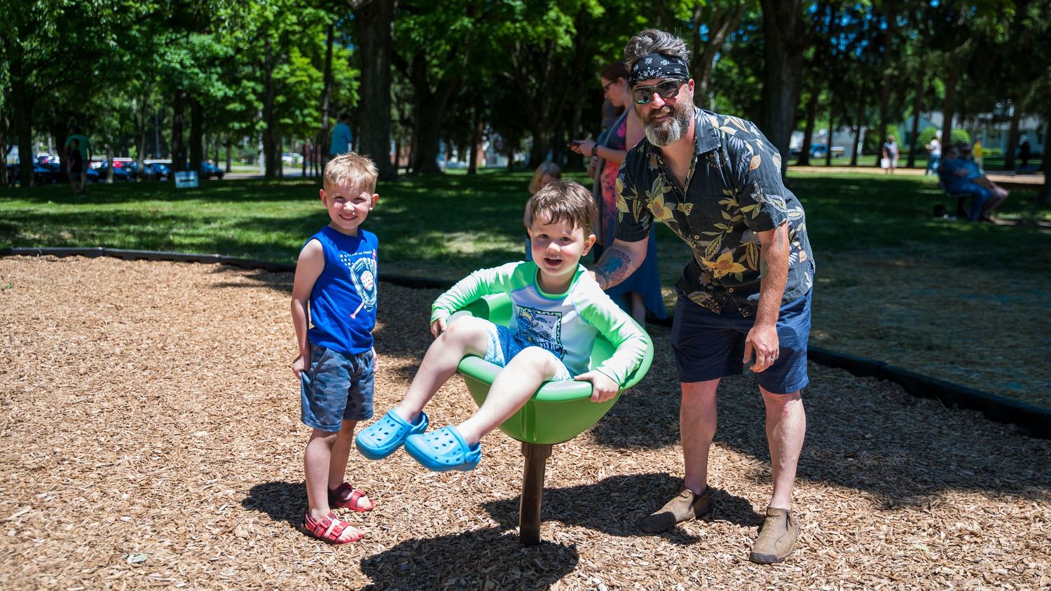 Two boys and a man enjoy a playground spinner at a sunny park with green trees and wood chip ground cover.
