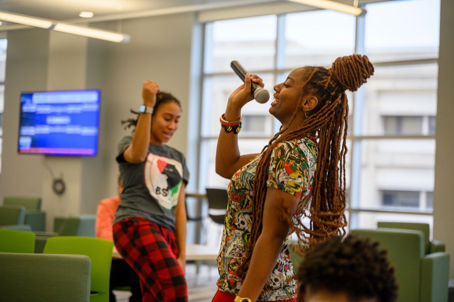 Young woman with braids sings into microphone while another person dances in a bright modern room.