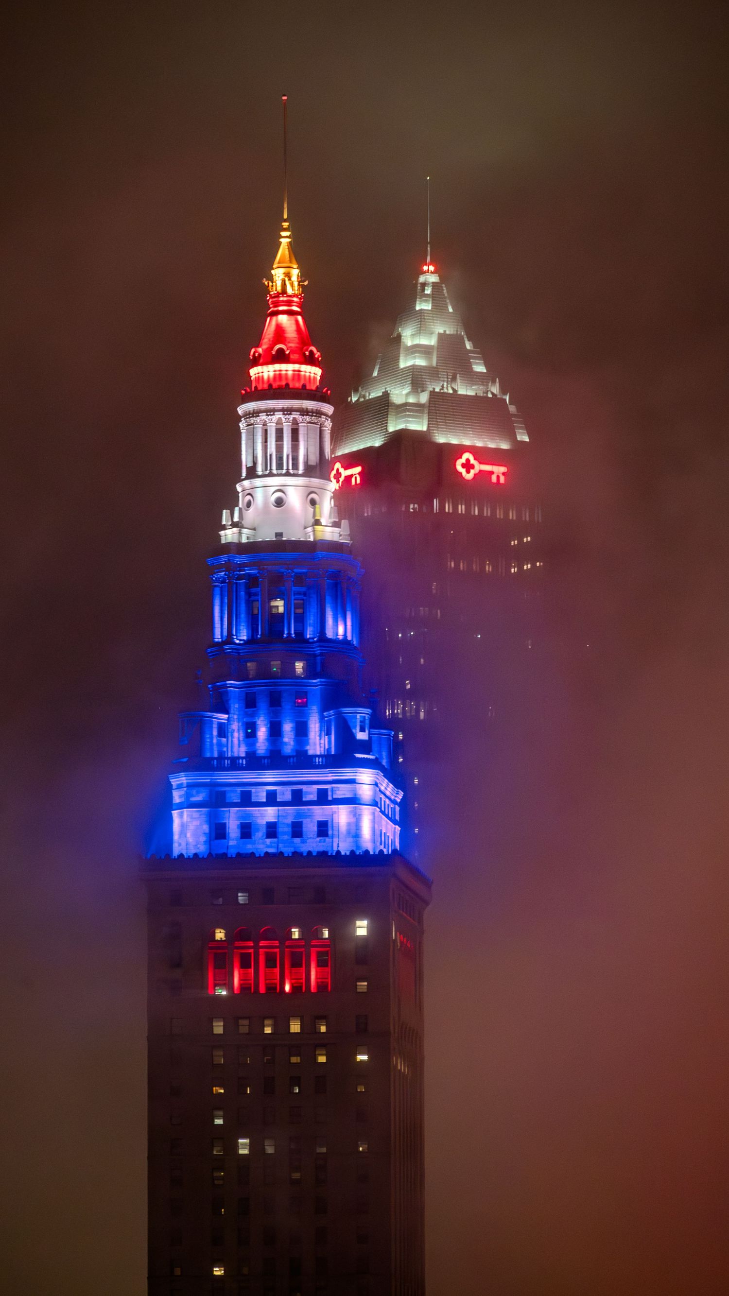 Terminal Tower in Cleveland illuminated in red, white, and blue lights surrounded by dramatic foggy night sky.