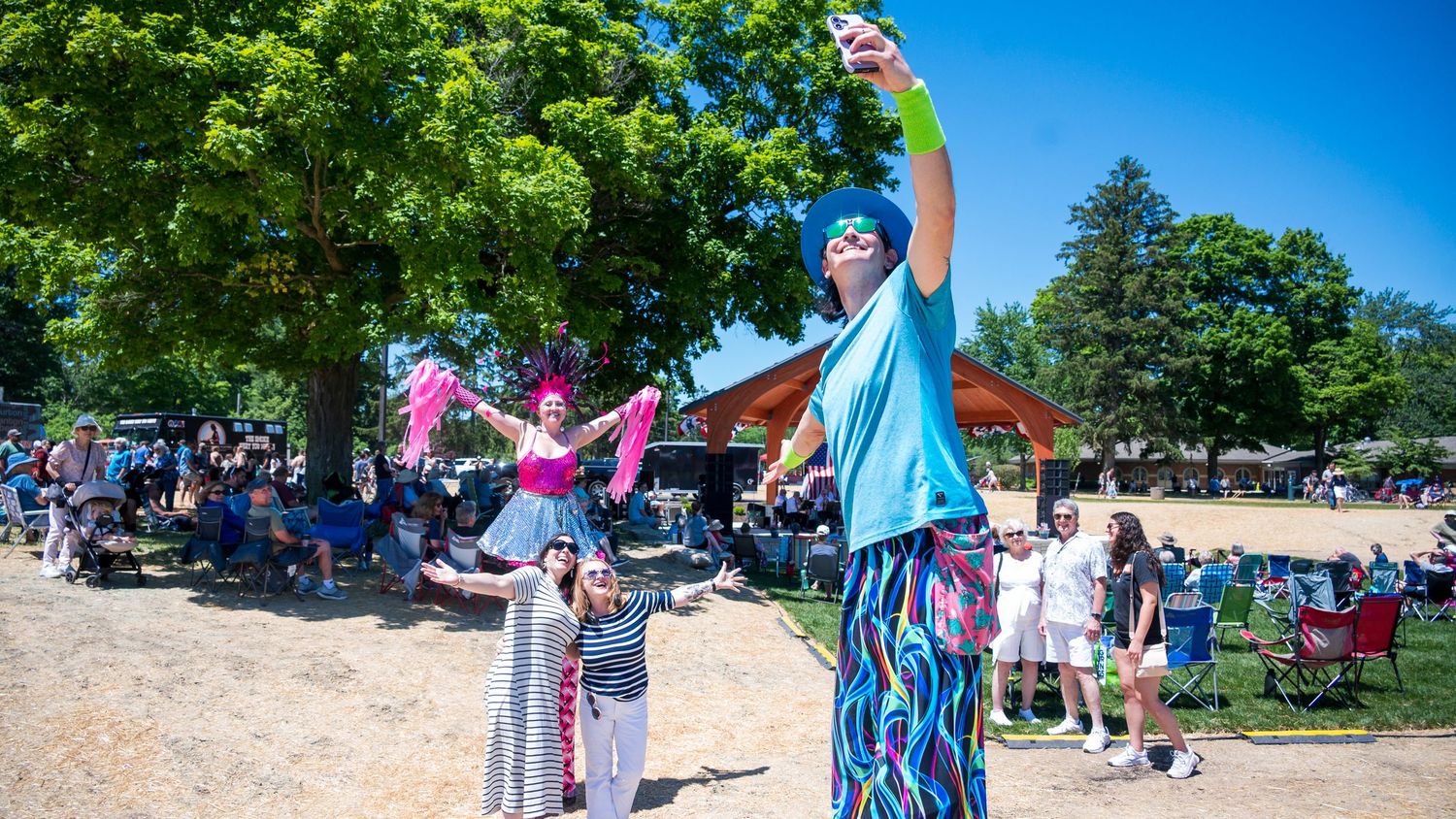 Performers in colorful costumes entertain crowds at an outdoor summer festival in a sunny park.