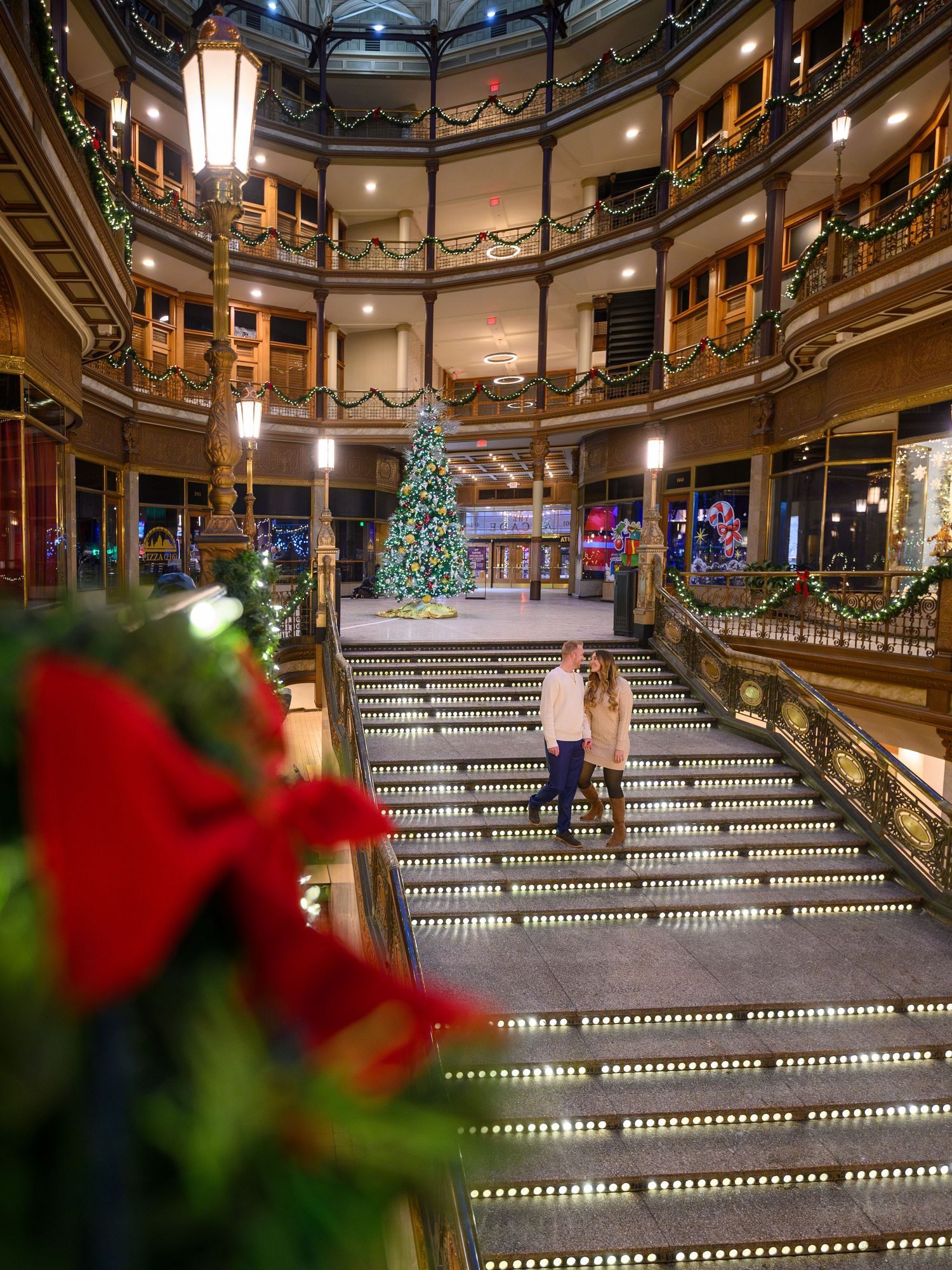 Couple stands on candlelit stairs in decorated holiday atrium with Christmas tree and garland.