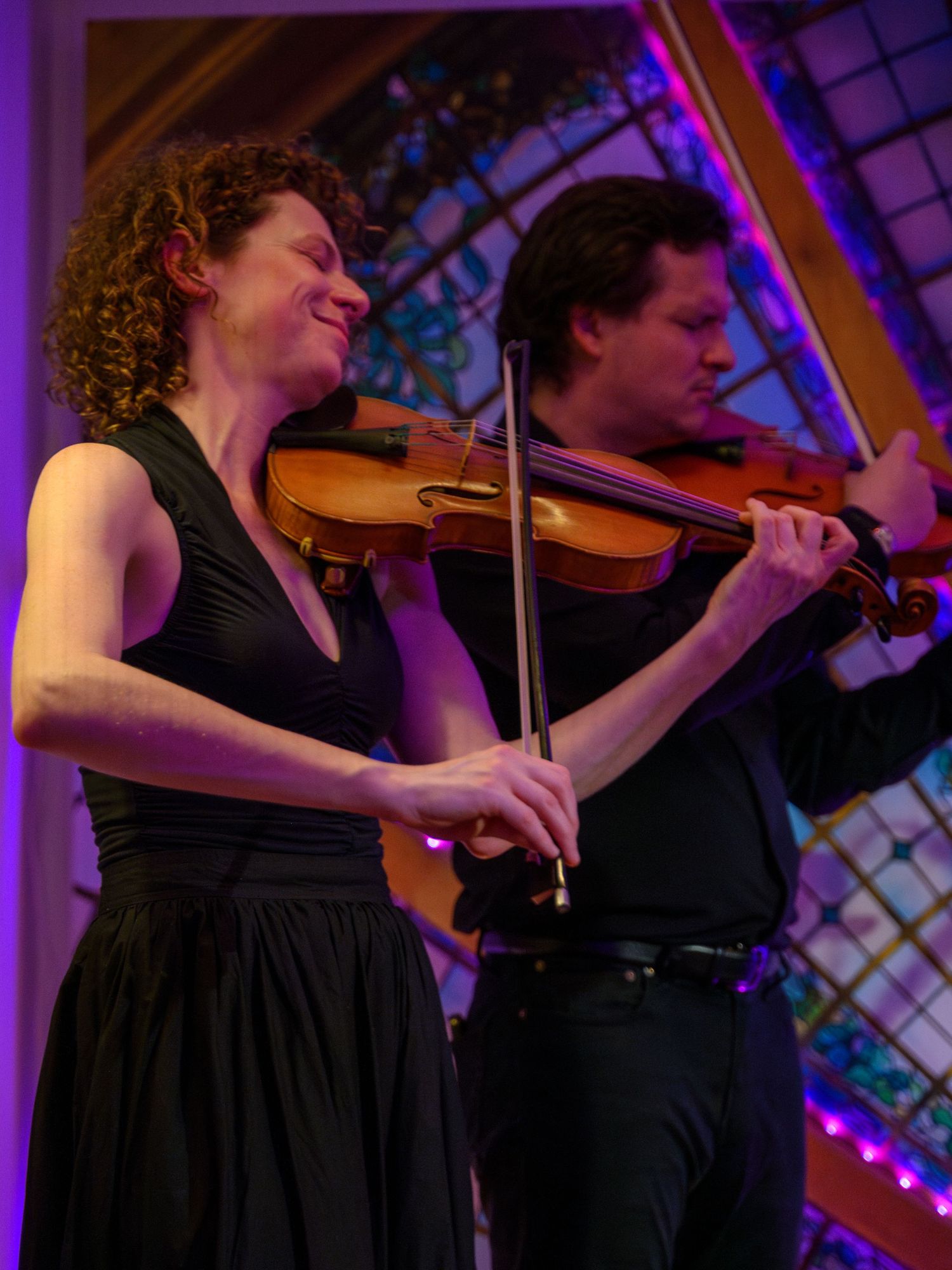 Two violinists perform in formal attire before colorful stained glass windows.
