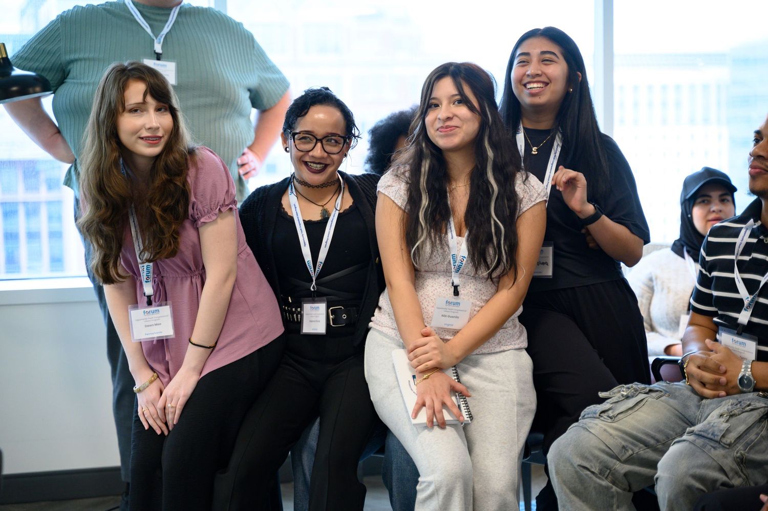 Four young women wearing lanyards smile and pose together at an indoor event or conference.