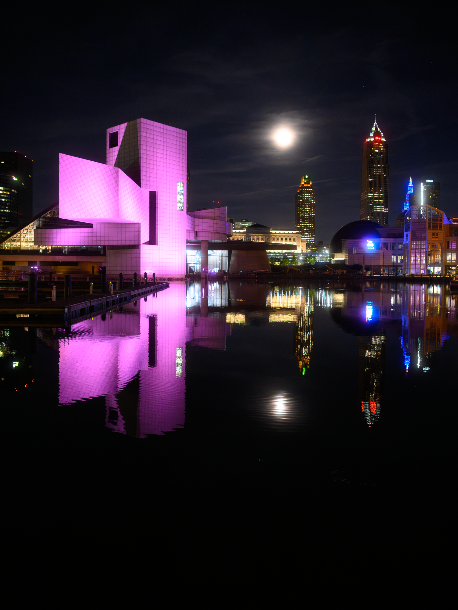 Rock and Roll Hall of Fame illuminated in pink light reflecting on water at night with full moon in Cleveland skyline.