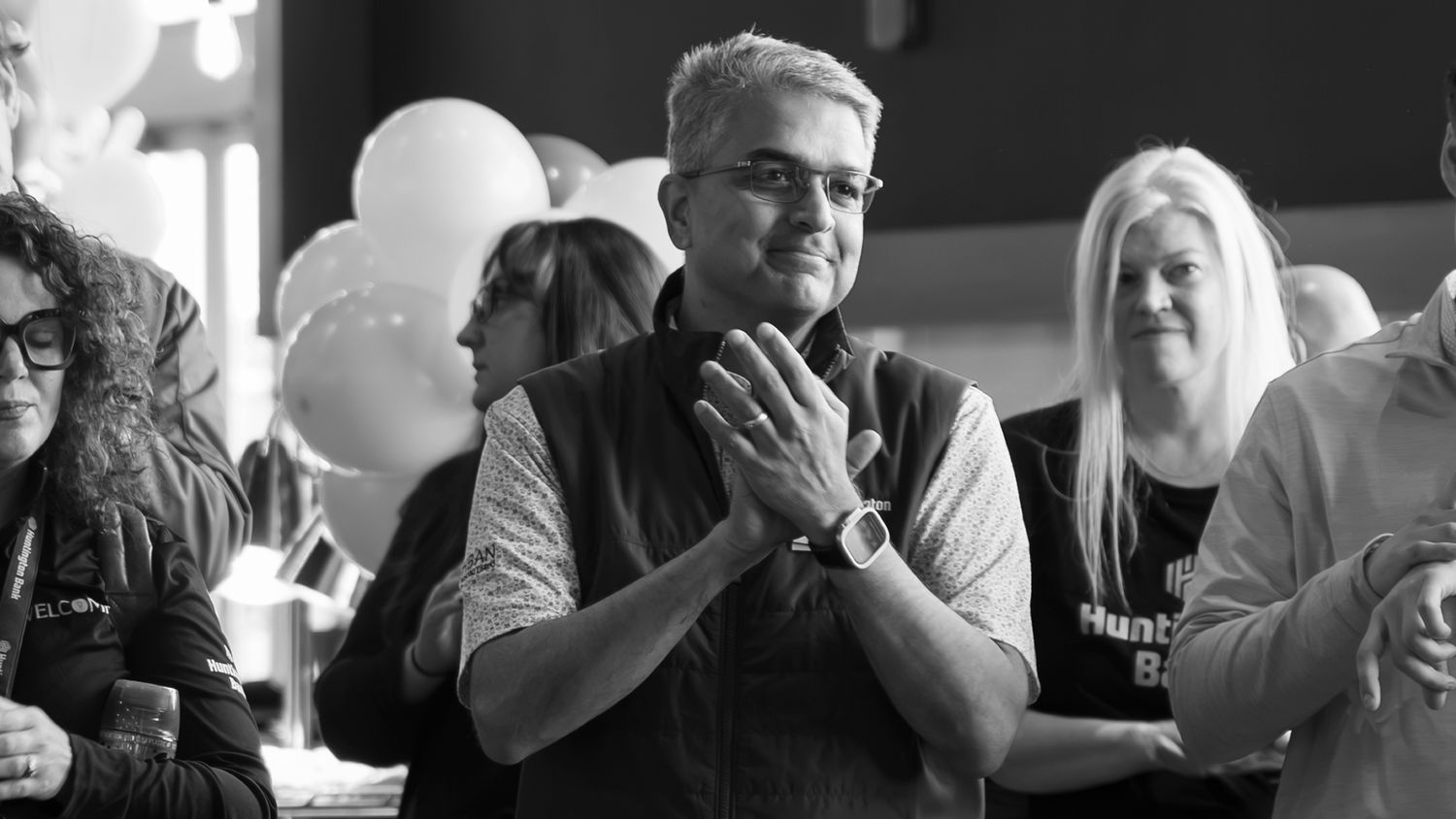 Black and white photo of man clapping at corporate celebration with balloons in background.