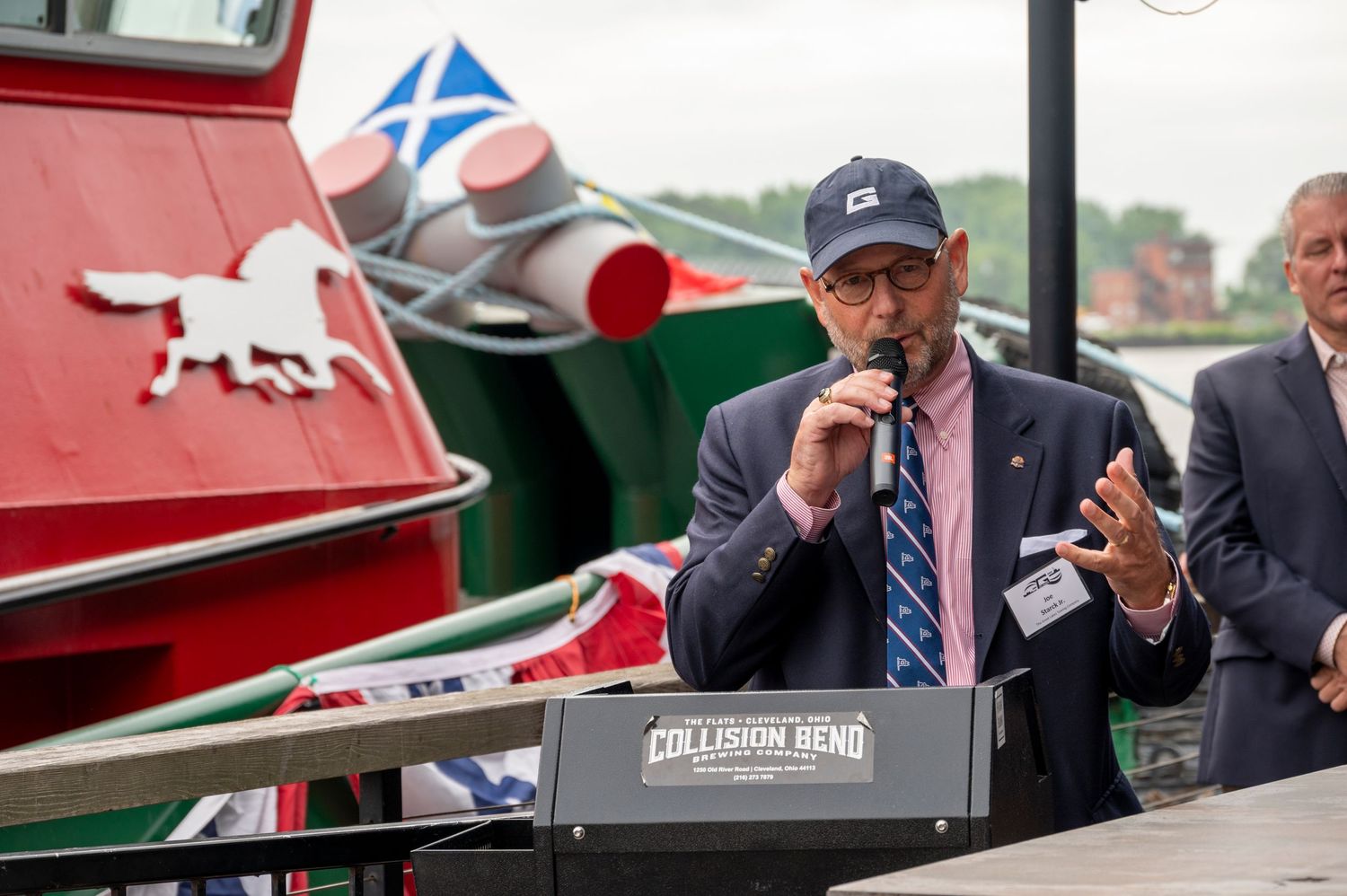 A man in a navy cap speaks into a microphone at an outdoor waterfront event near a colorful tugboat.