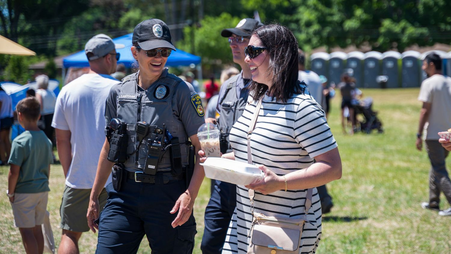 A police officer in uniform walks and talks with a civilian woman at an outdoor community event on a sunny day.