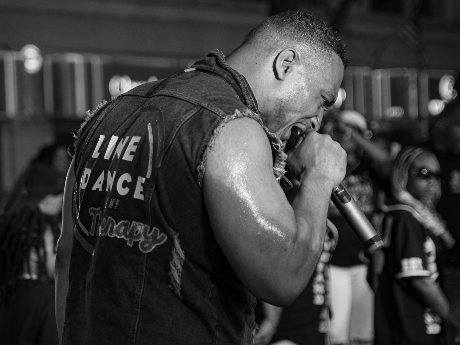 Black and white photo of a muscular man in a Line Dance vest singing into a microphone at an outdoor event.