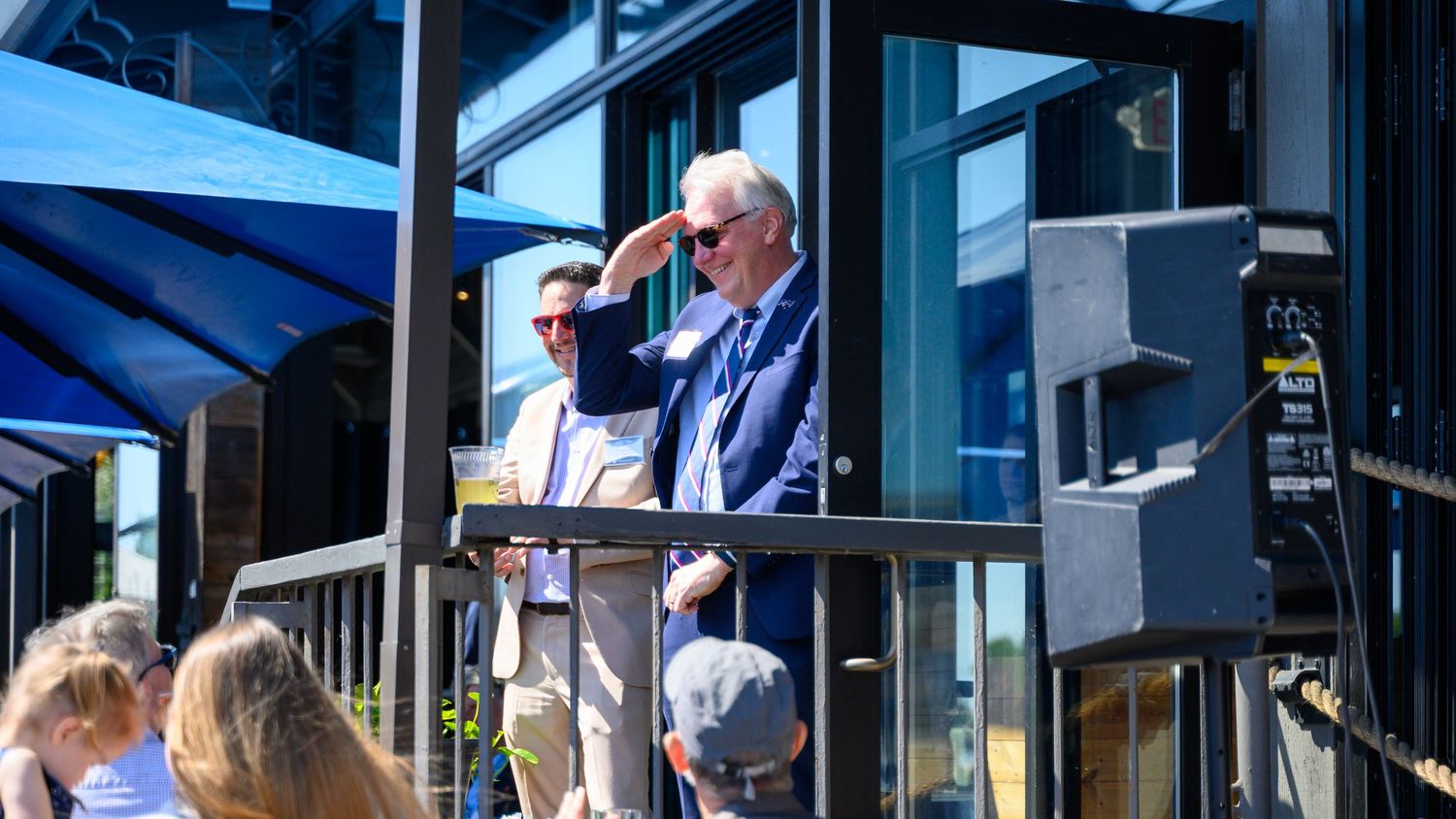 Two men stand at an outdoor podium on a sunny day, one shielding his eyes from the sun in a blue suit.