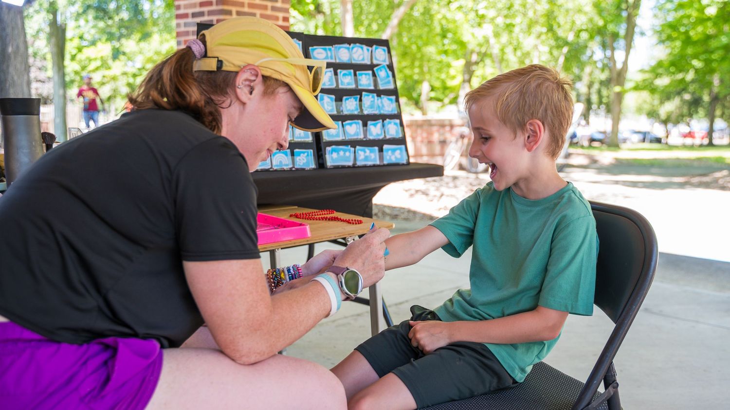 A woman in a yellow cap applies face paint to a smiling young boy at an outdoor summer event.