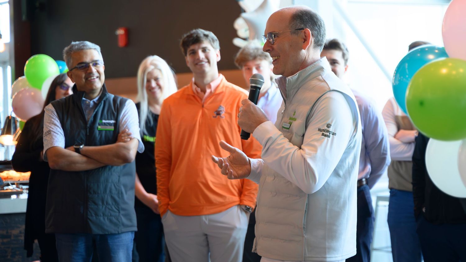 Man speaking with microphone at colorful balloon-decorated corporate team celebration event.