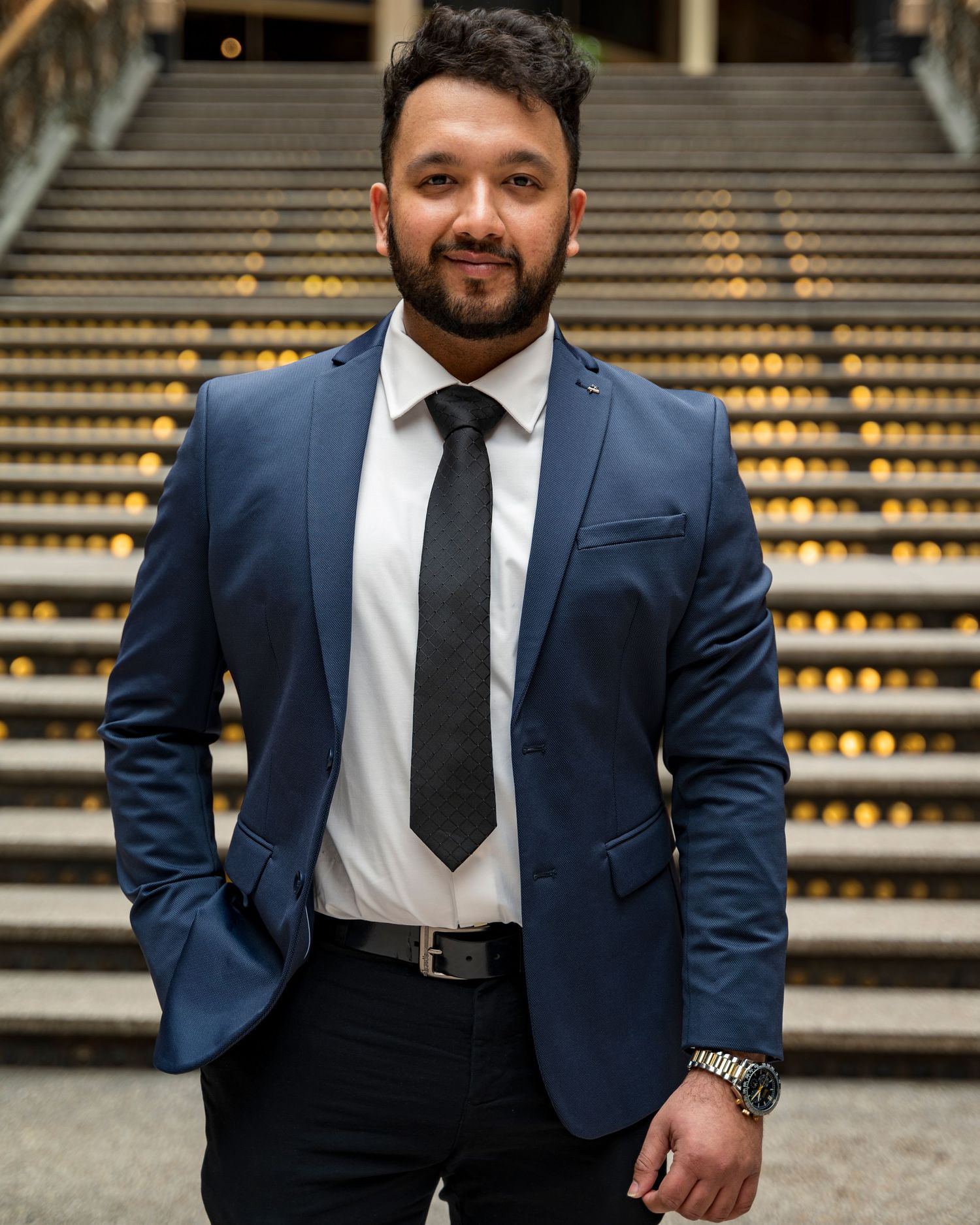 Professional man in navy blue suit and black tie standing on decorative staircase.