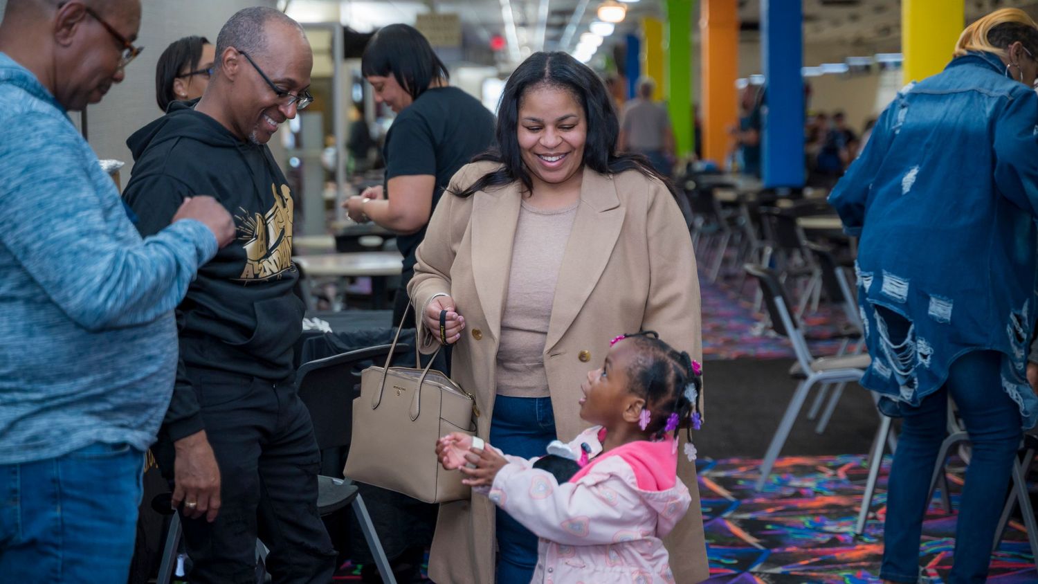 A woman in a beige blazer smiles at a young girl in a pink outfit at an indoor community event.