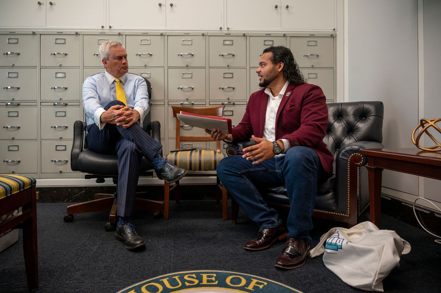 Two men in business attire sit in chairs having a conversation in an office with a House of Representatives seal.